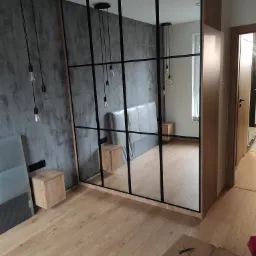 Nowoczesna szafa wnękowa z czarnymi profilami szklanymi, widoczna w sypialni z szarą ścianą o fakturze betonu i drewnianą podłogą; karton na podłodze sugeruje świeży montaż.