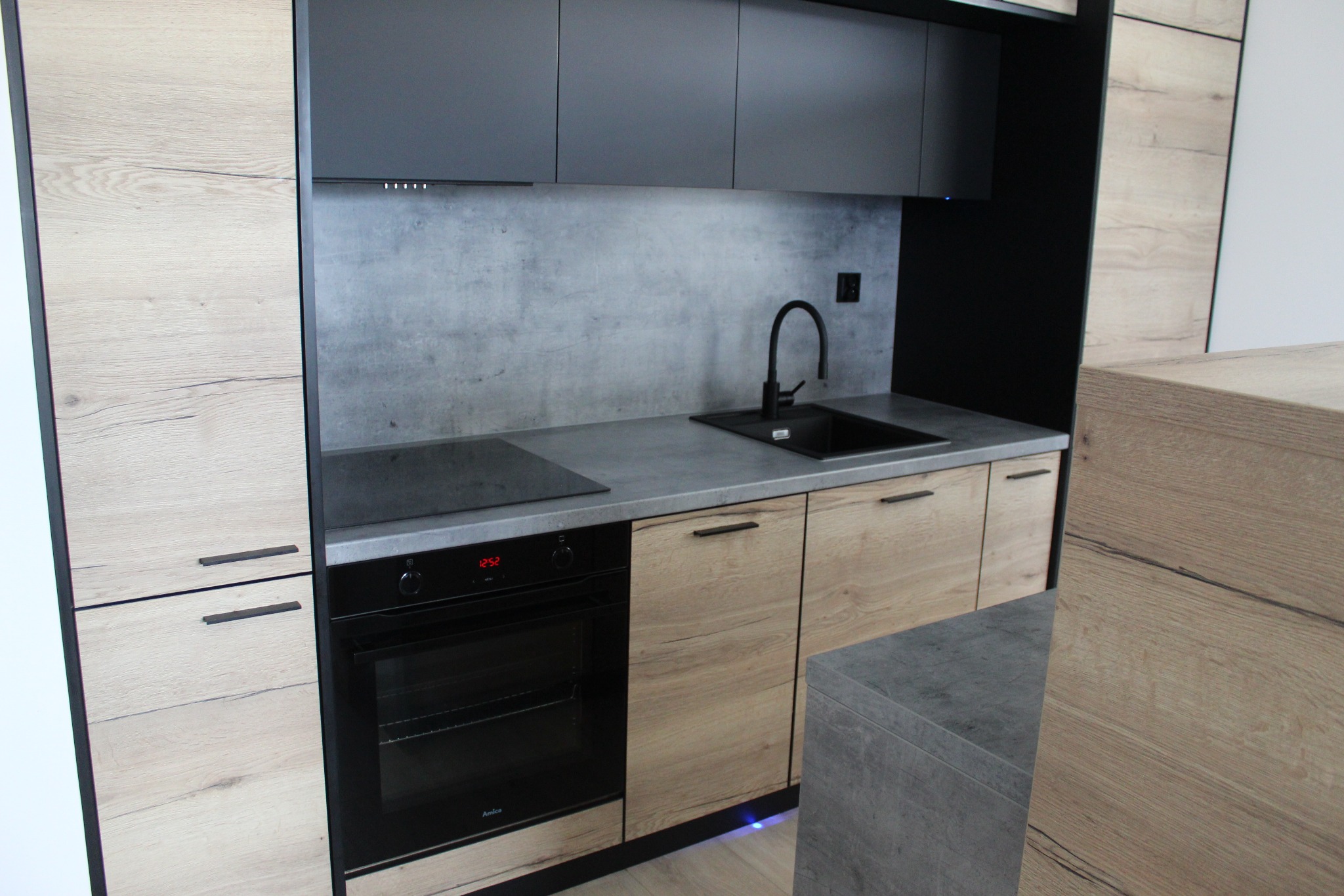 Nowoczesna kuchnia z drewnianymi frontami szafek dolnych i ciemnoszarymi górnymi, szary blat i ściana nad blatem, czarny zlew i bateria, piekarnik Amica.