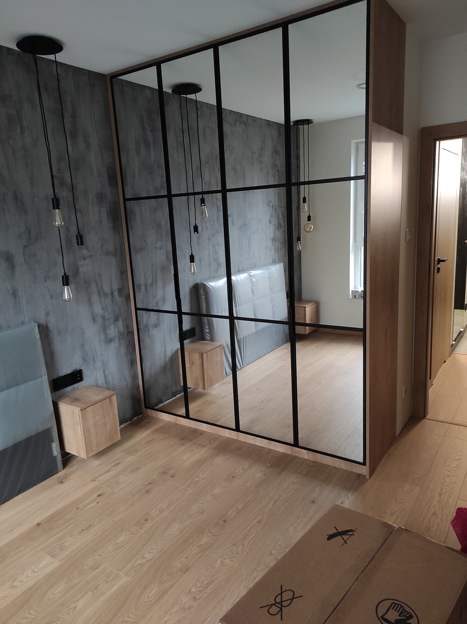 Nowoczesna szafa wnękowa z czarnymi profilami szklanymi, widoczna w sypialni z szarą ścianą o fakturze betonu i drewnianą podłogą; karton na podłodze sugeruje świeży montaż.