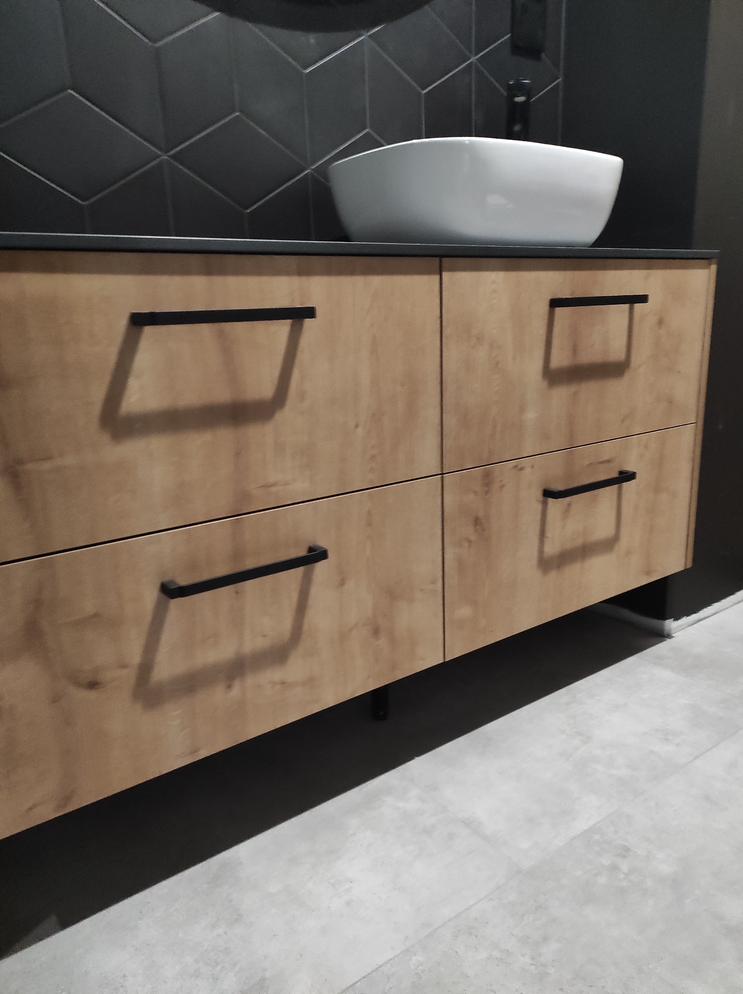 Łazienkowa szafka pod umywalkę z drewnopodobnym frontem, czarne uchwyty, biała umywalka nablatowa, tło z czarnych płytek w geometryczny wzór.