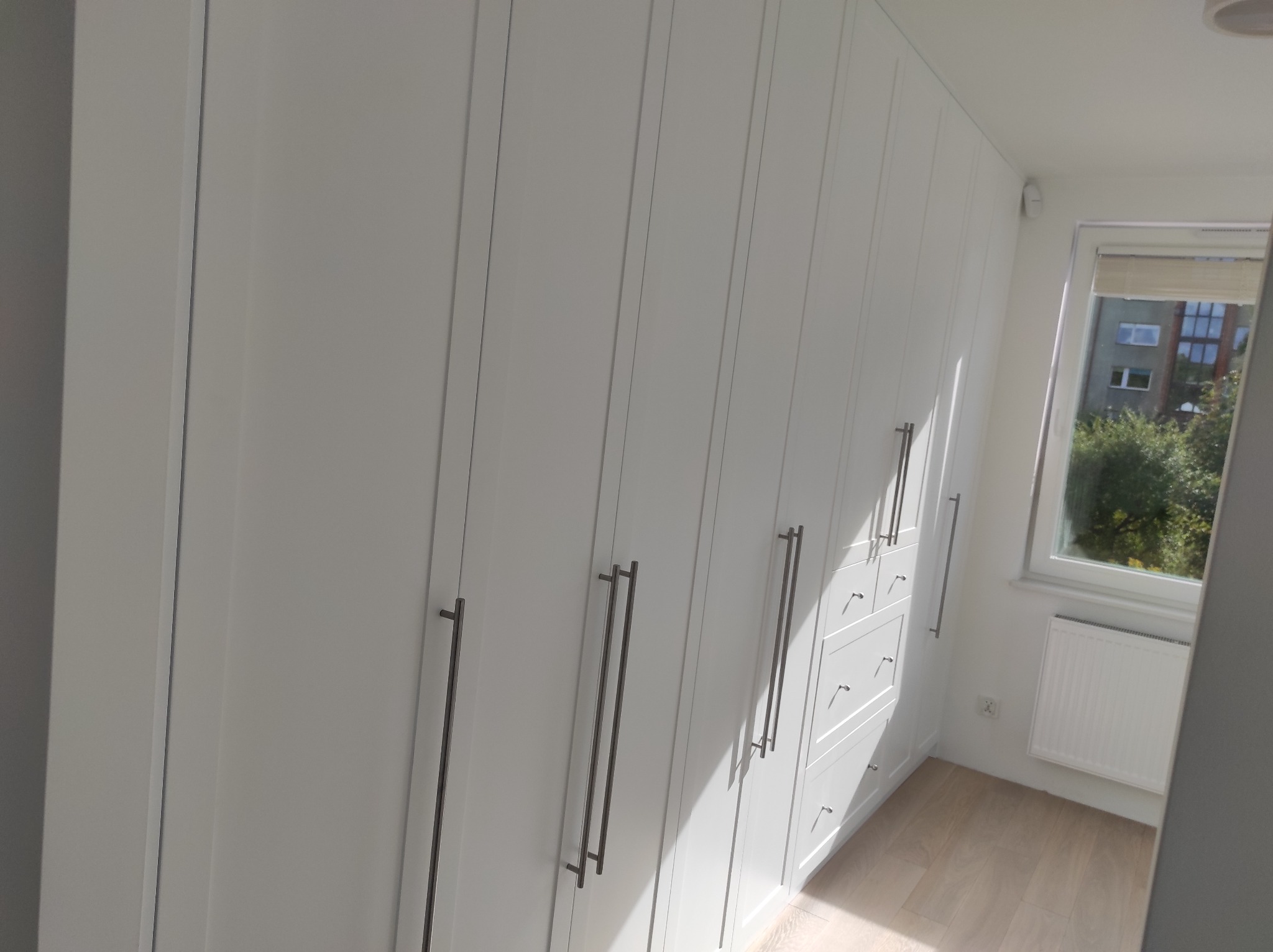 Biała szafa wnękowa z długimi, pionowymi uchwytami, oświetlona naturalnym światłem wpadającym przez okno z widokiem na drzewa.