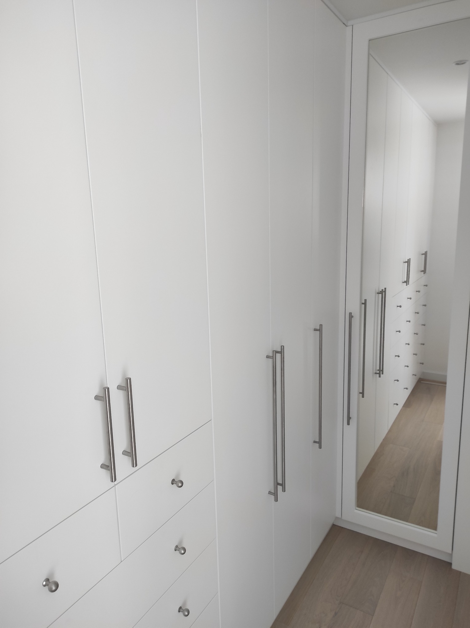 Biała szafa wnękowa z długimi, pionowymi uchwytami i lustrem na drzwiach, odbijającym wnętrze szafy z szufladami i półkami, na podłodze panele drewniane.