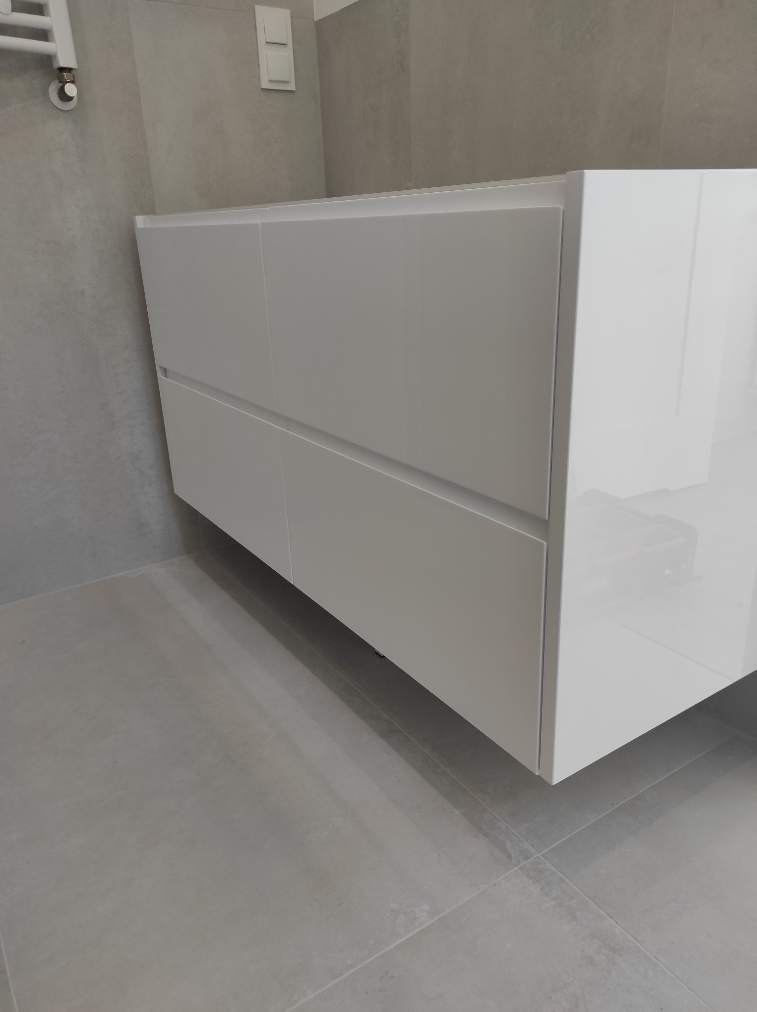 Biała, podwieszana szafka łazienkowa z dwiema szufladami, z połyskiem, na tle szarej ściany i podłogi z płytek ceramicznych.