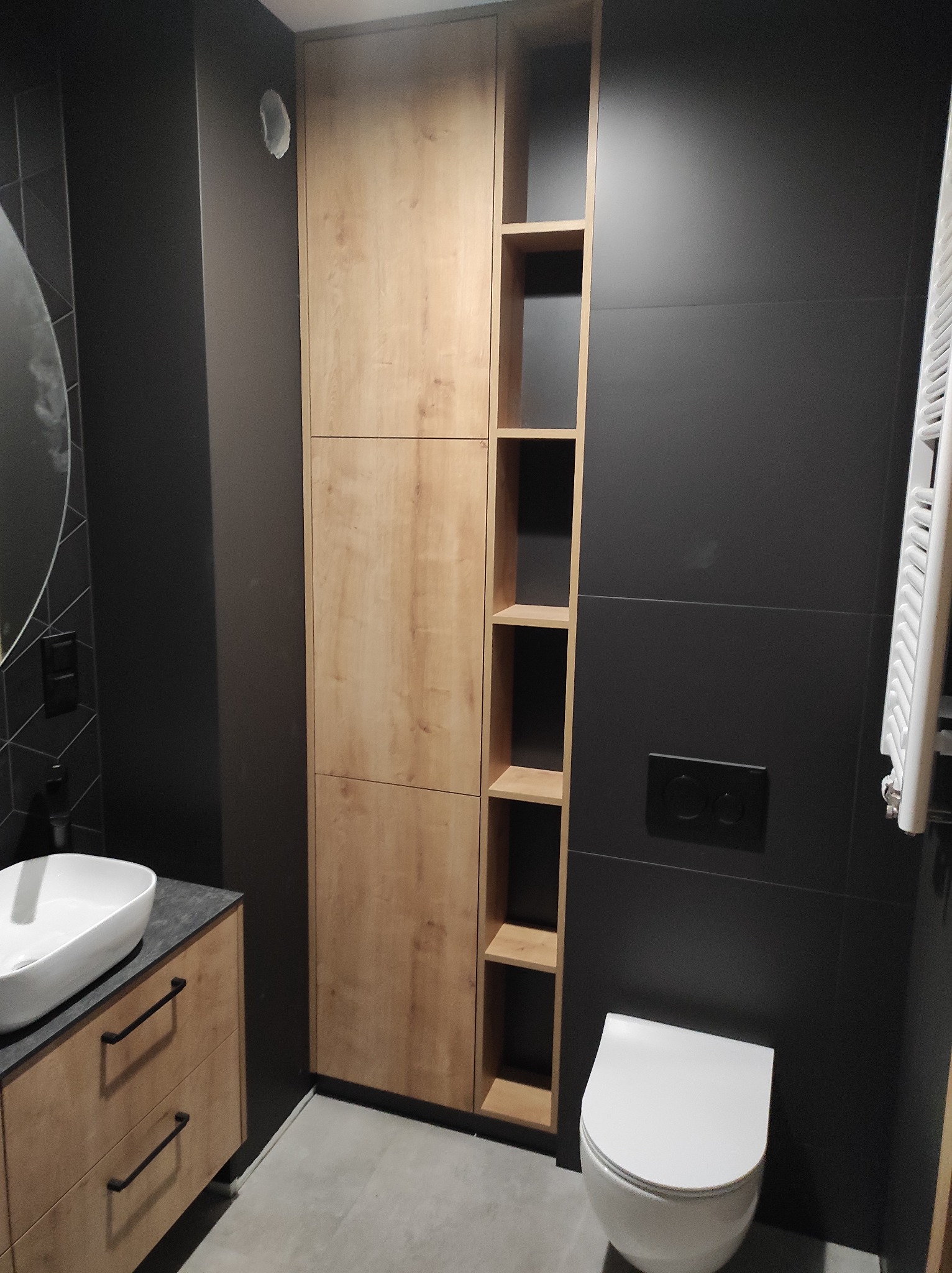 Nowoczesna łazienka z drewnianą szafką pod umywalką, wysoką szafą z otwartymi półkami i czarnymi płytkami na ścianach, oświetlona neutralnym światłem.