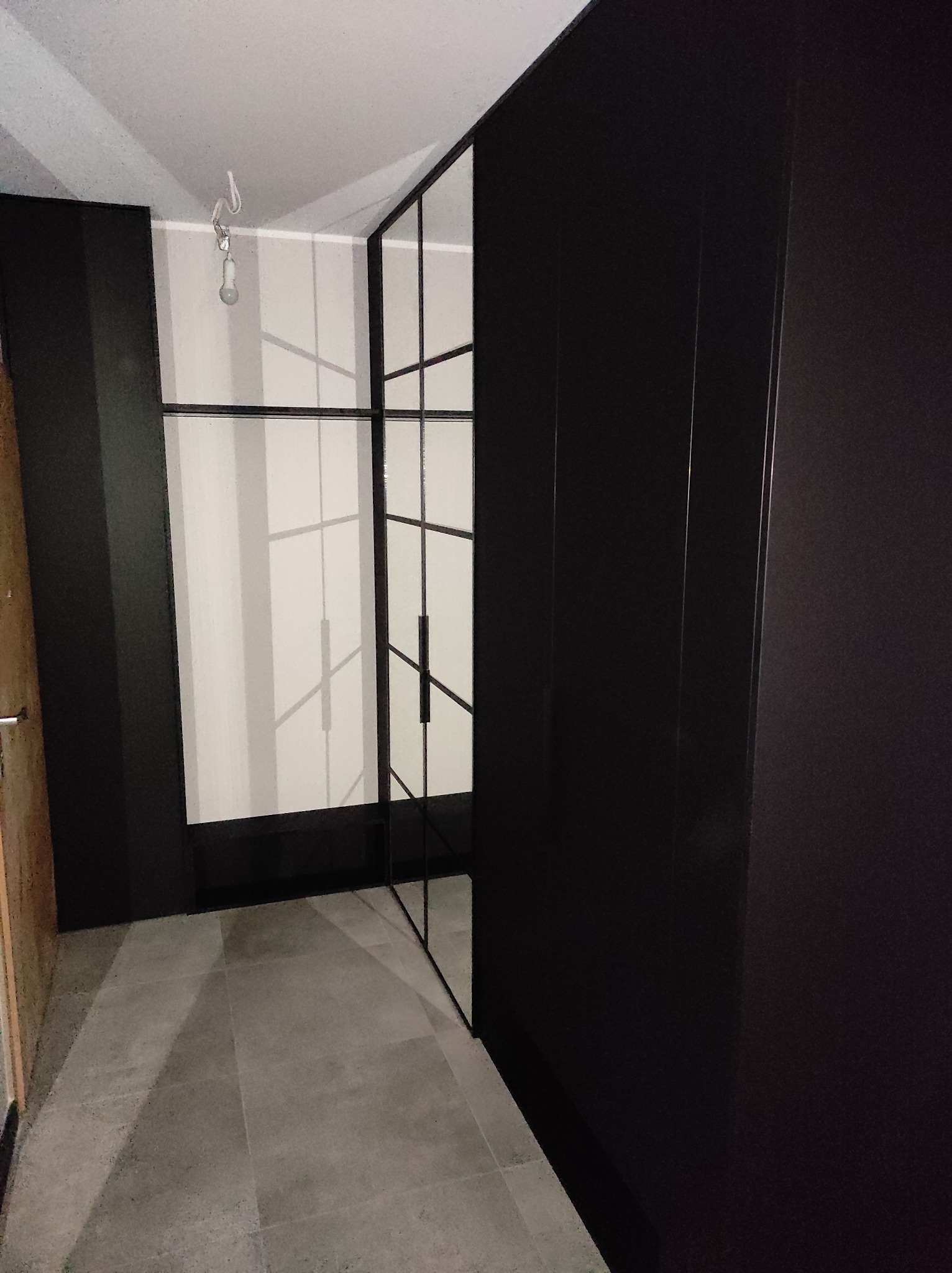 Nowoczesna szafa wnękowa z czarnymi frontami i szklanymi drzwiami z metalowymi szprosami, widok na szarą podłogę i surowe oświetlenie sufitowe.