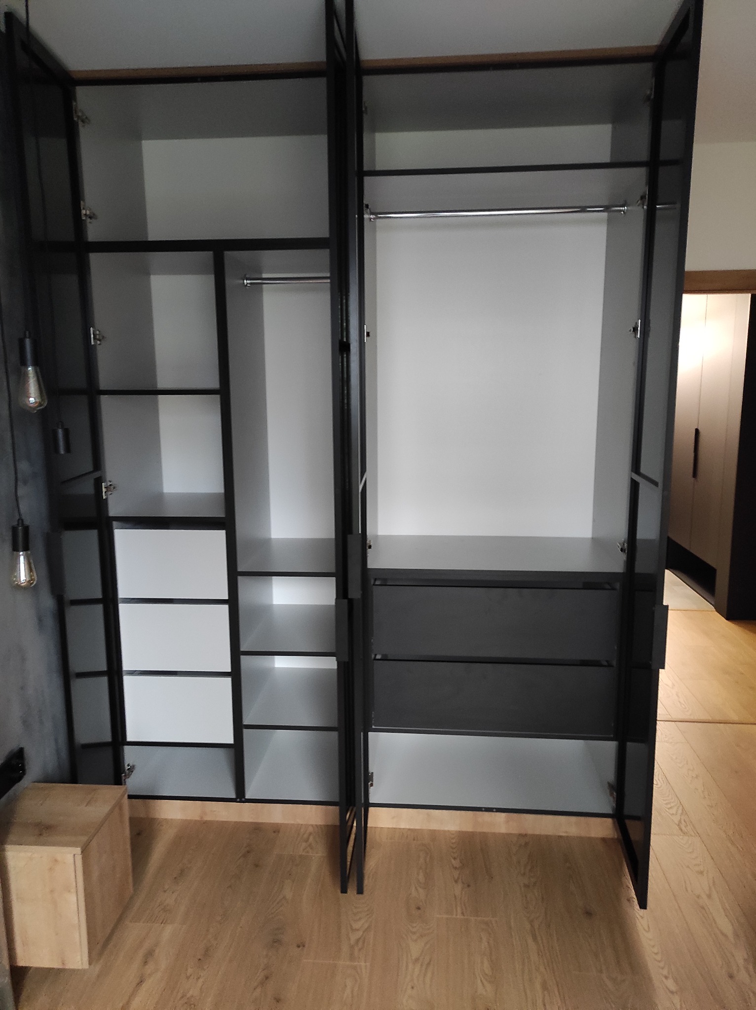 Otwarta szafa wnękowa z czarną ramą, białymi półkami i szufladami, widoczne detale wykończenia i oświetlenie boczne z żarówkami edisona.