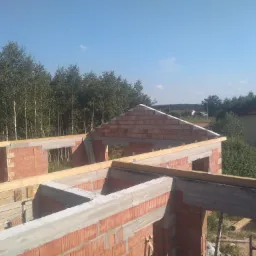 Wznoszenie ścian z czerwonej cegły i betonowych nadproży domu jednorodzinnego w Radomiu, widok z góry na konstrukcję w stanie surowym otwartym, z lasem w tle.