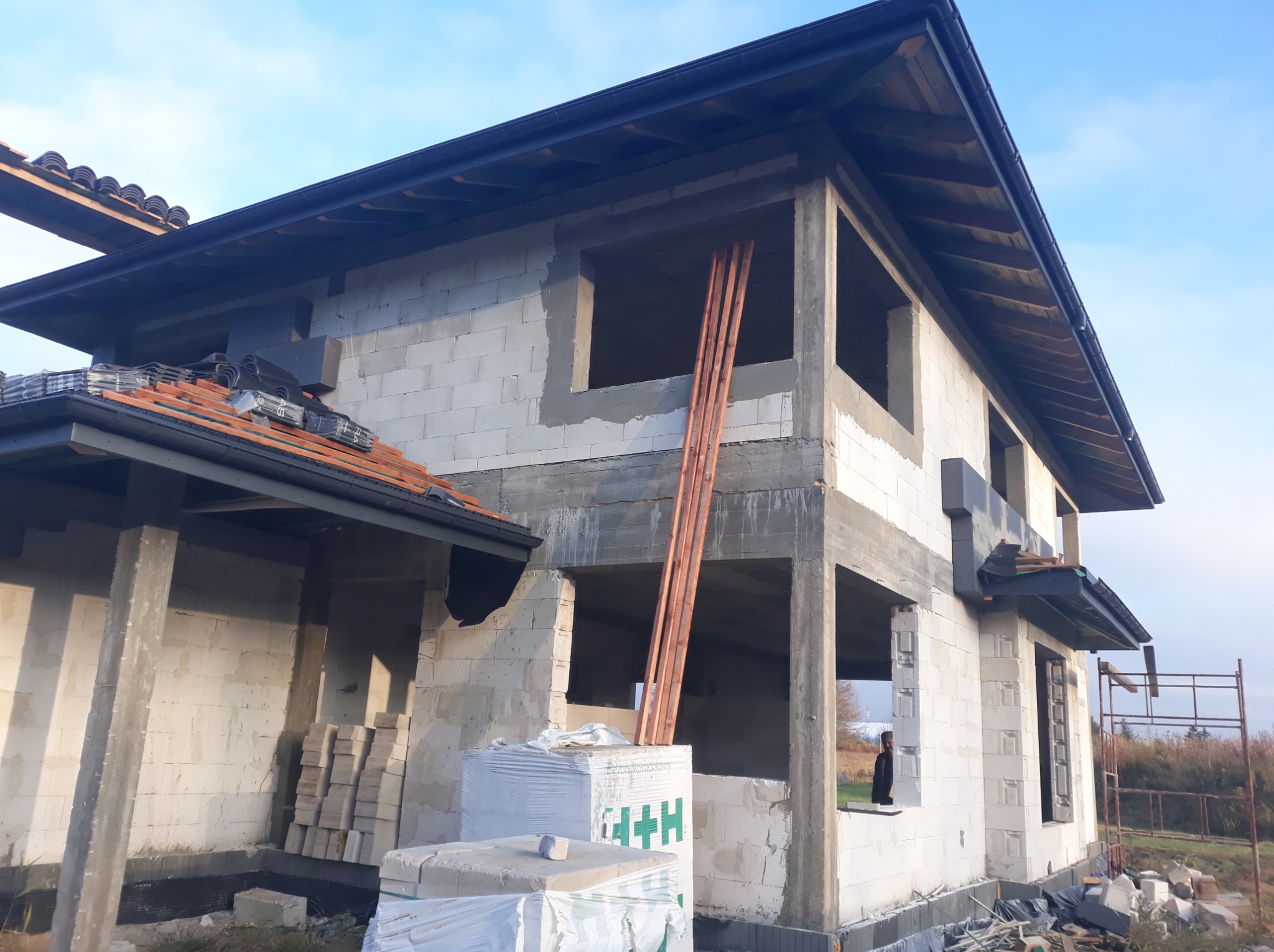 Dwukondygnacyjny dom w stanie surowym otwartym, zbudowany z białych bloczków, z częściowo położonym dachem z dachówki ceramicznej, widoczne drewniane łaty opierające się o ścianę.