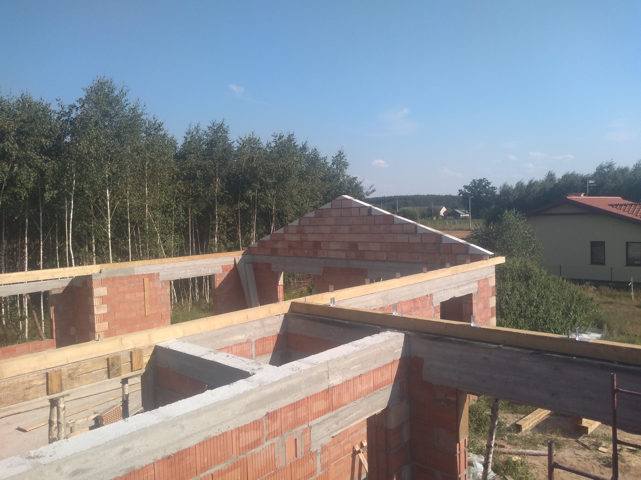 Wznoszenie ścian z czerwonej cegły i betonowych nadproży domu jednorodzinnego w Radomiu, widok z góry na konstrukcję w stanie surowym otwartym, z lasem w tle.