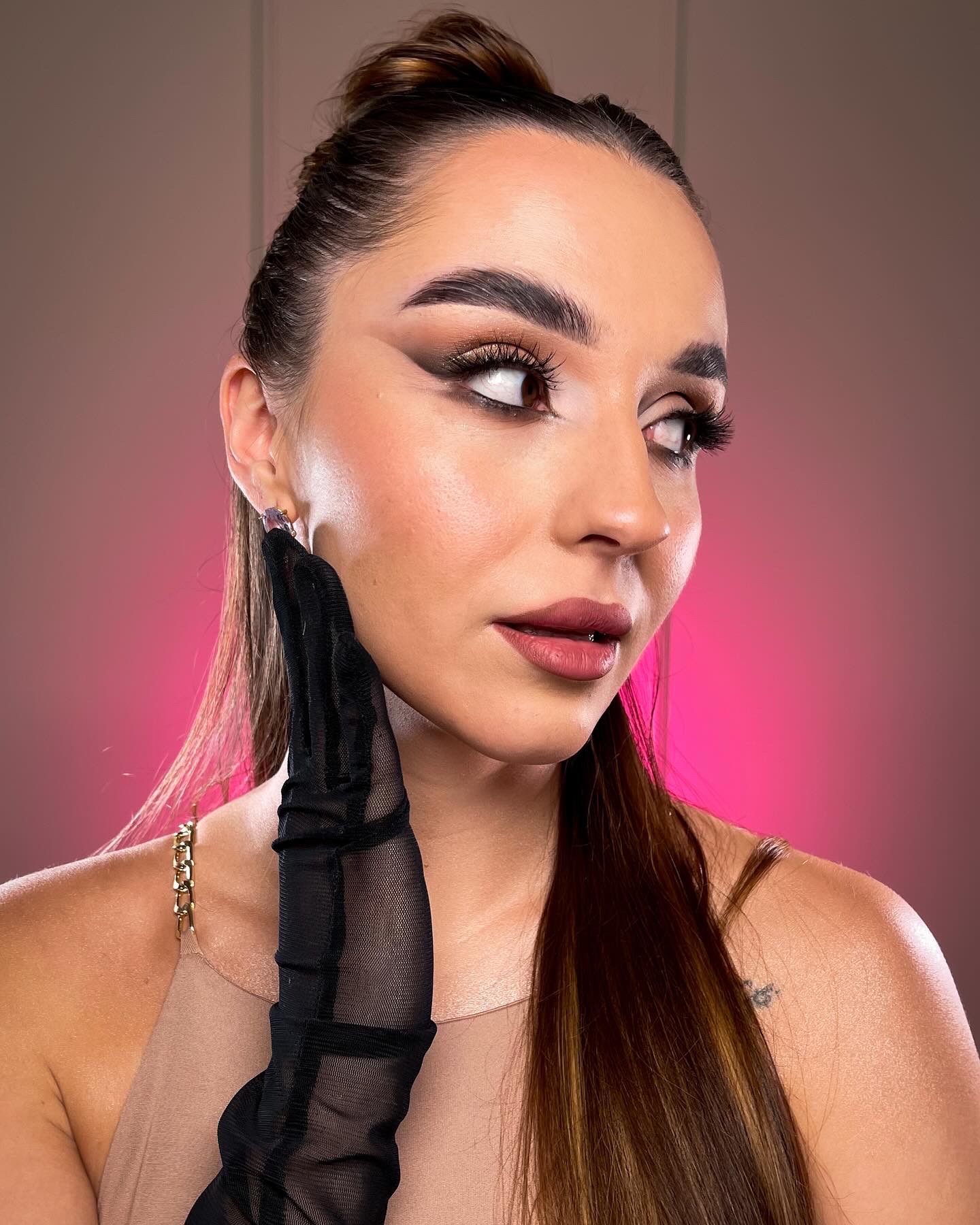 Portret kobiety z wyrazistym makijażem oczu w stylu cat-eye, z czarną, siateczkową rękawiczką dotykającą policzka, z upiętymi włosami i delikatną biżuterią na tle z gradientem różu i beżu.