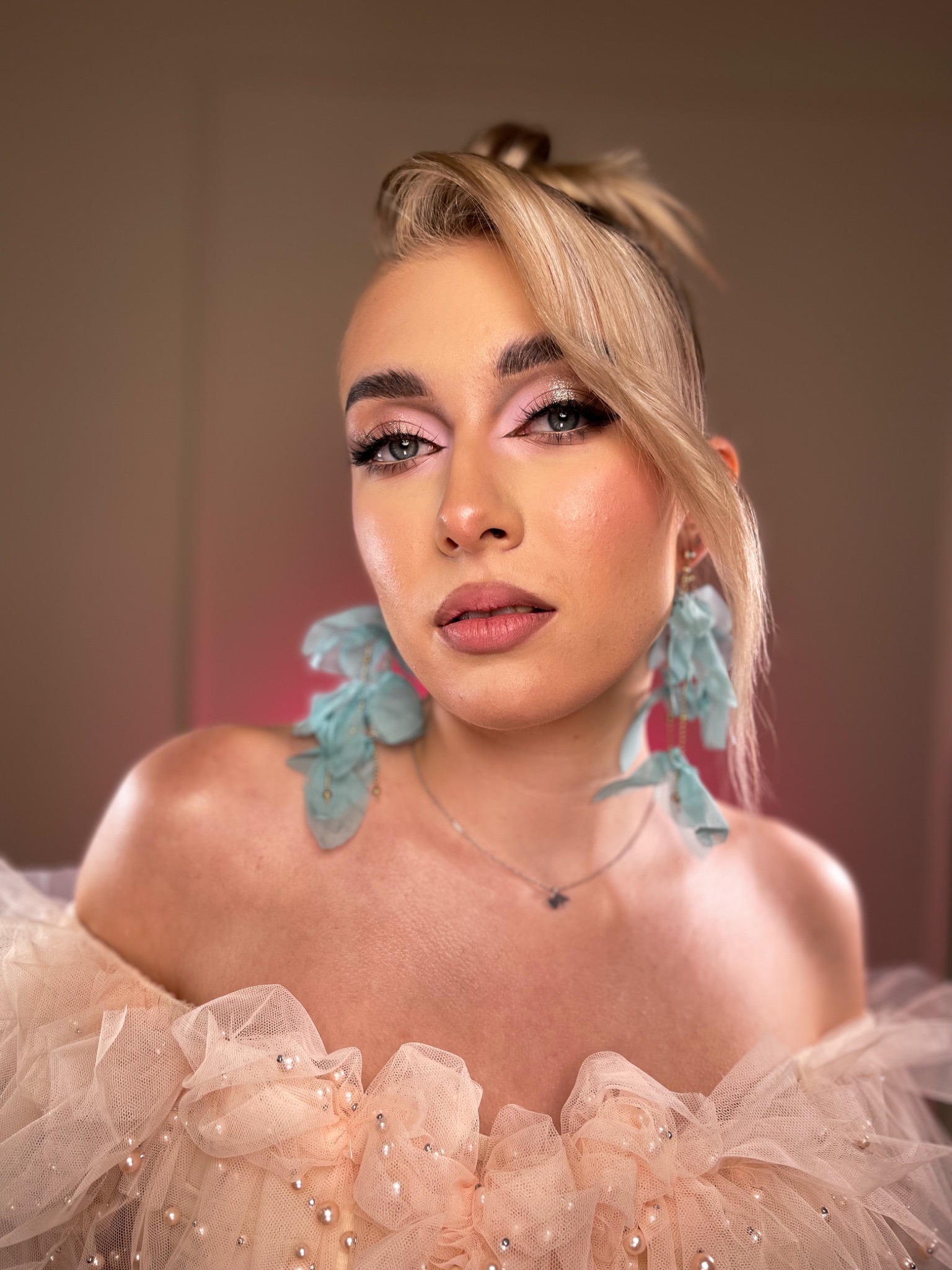 Portret kobiety z blond włosami upiętymi w kok, delikatny różowy makijaż oczu i ust, duże turkusowe kolczyki z tiulu i sukienka z falbanami i perłami.