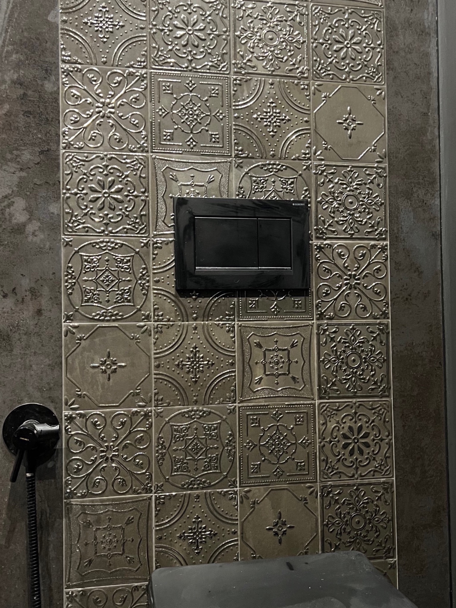 Ściana w łazience wykończona dekoracyjnymi, tłoczonymi płytkami ceramicznymi w odcieniach szarości, z zamontowanym czarnym przyciskiem spłuczki i fragmentem bidetki.