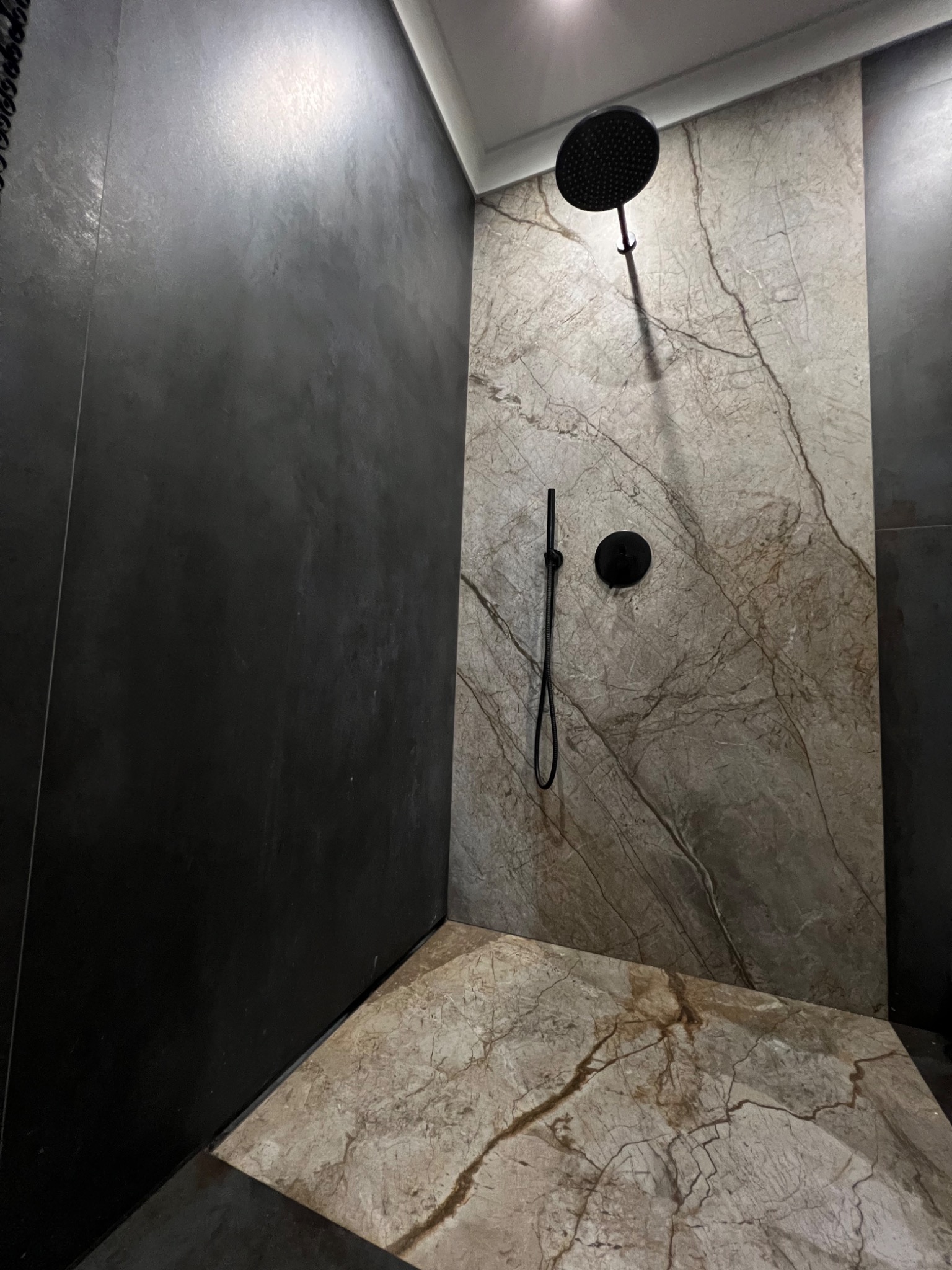 Nowoczesna kabina prysznicowa z czarną armaturą i płytkami imitującymi marmur na ścianach i podłodze, kontrastująca z ciemnoszarą ścianą obok.