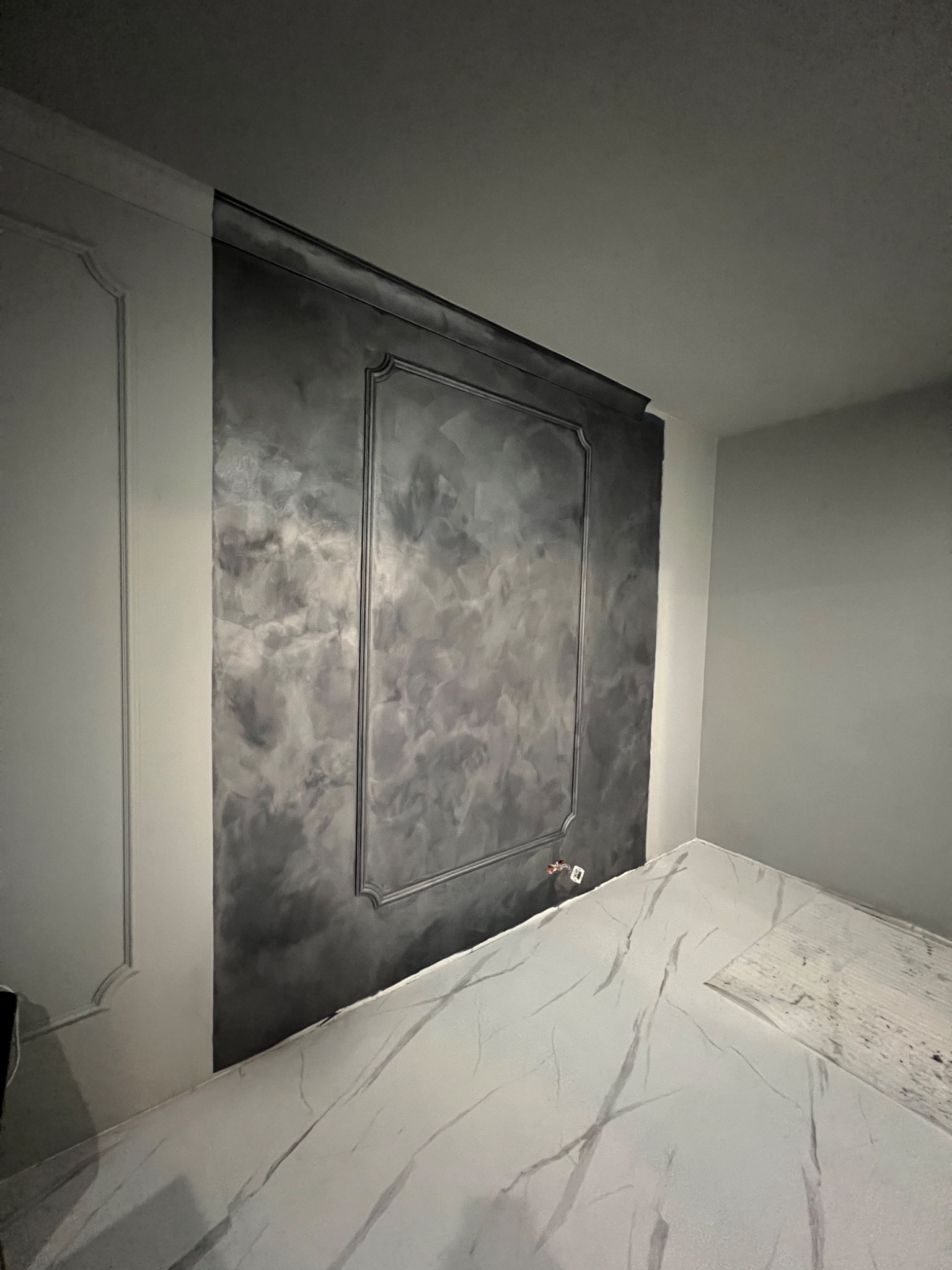 Ściana z dekoracyjną, szarą gładzią w ramie, marmurowa podłoga z białymi żyłami, widoczne gniazdko elektryczne, fragment sztukaterii.
