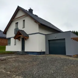 Nowoczesny dom jednorodzinny z białą elewacją, szarą podmurówką i garażem w kolorze grafitowym, widoczny z perspektywy podjazdu wysypanego żwirem.