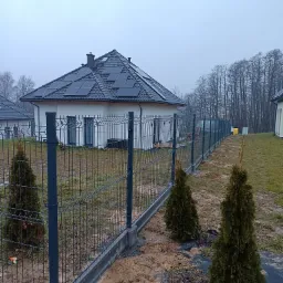 Nowoczesny dom jednorodzinny z panelami słonecznymi na dachu i ogrodzeniem panelowym z podmurówką, widoczne tuje i błotniste podłoże.