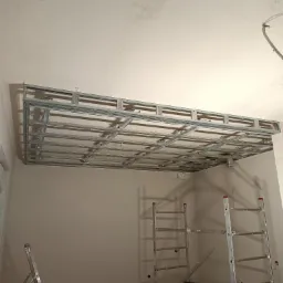 Metalowa konstrukcja sufitu podwieszanego z płyt karton-gips, widoczna na tle surowego sufitu i ścian w trakcie montażu, z użyciem drabiny i rusztowania.