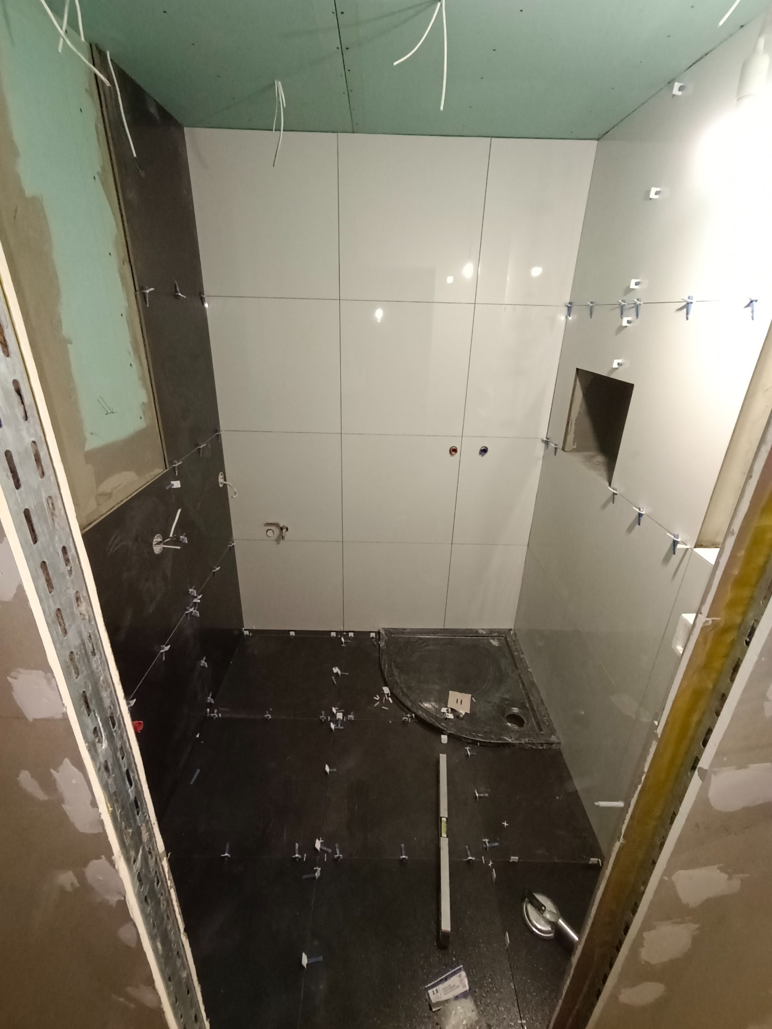Wnętrze łazienki w trakcie prac wykończeniowych, widoczne ciemne płytki podłogowe z krzyżykami dystansowymi, jasne płytki ścienne częściowo ułożone, poziomica, oraz elementy montażowe profili.