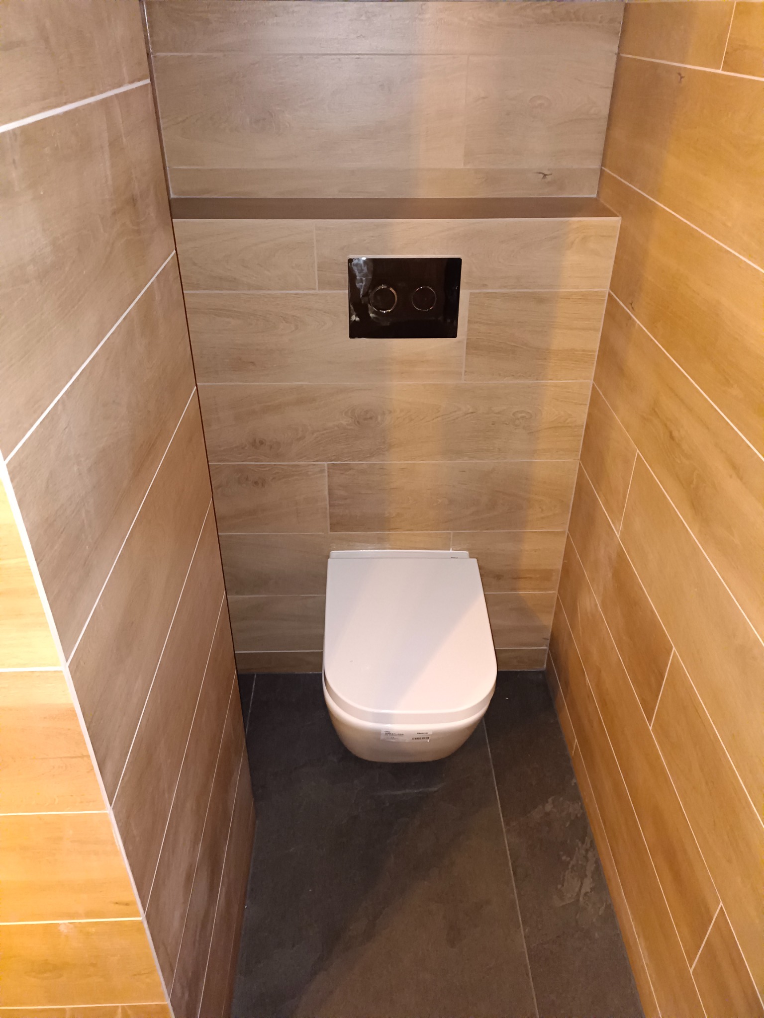 Nowoczesna toaleta podwieszana z białym siedziskiem, wkomponowana w przestrzeń wyłożoną drewnopodobnymi płytkami w odcieniach beżu i brązu, z czarnym panelem spłukującym i ciemną podłogą.