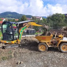 Żółta mini koparka JCB ładuje ziemię do wywrotki na gąsienicach; w tle widoczne domy, drzewa i góry.