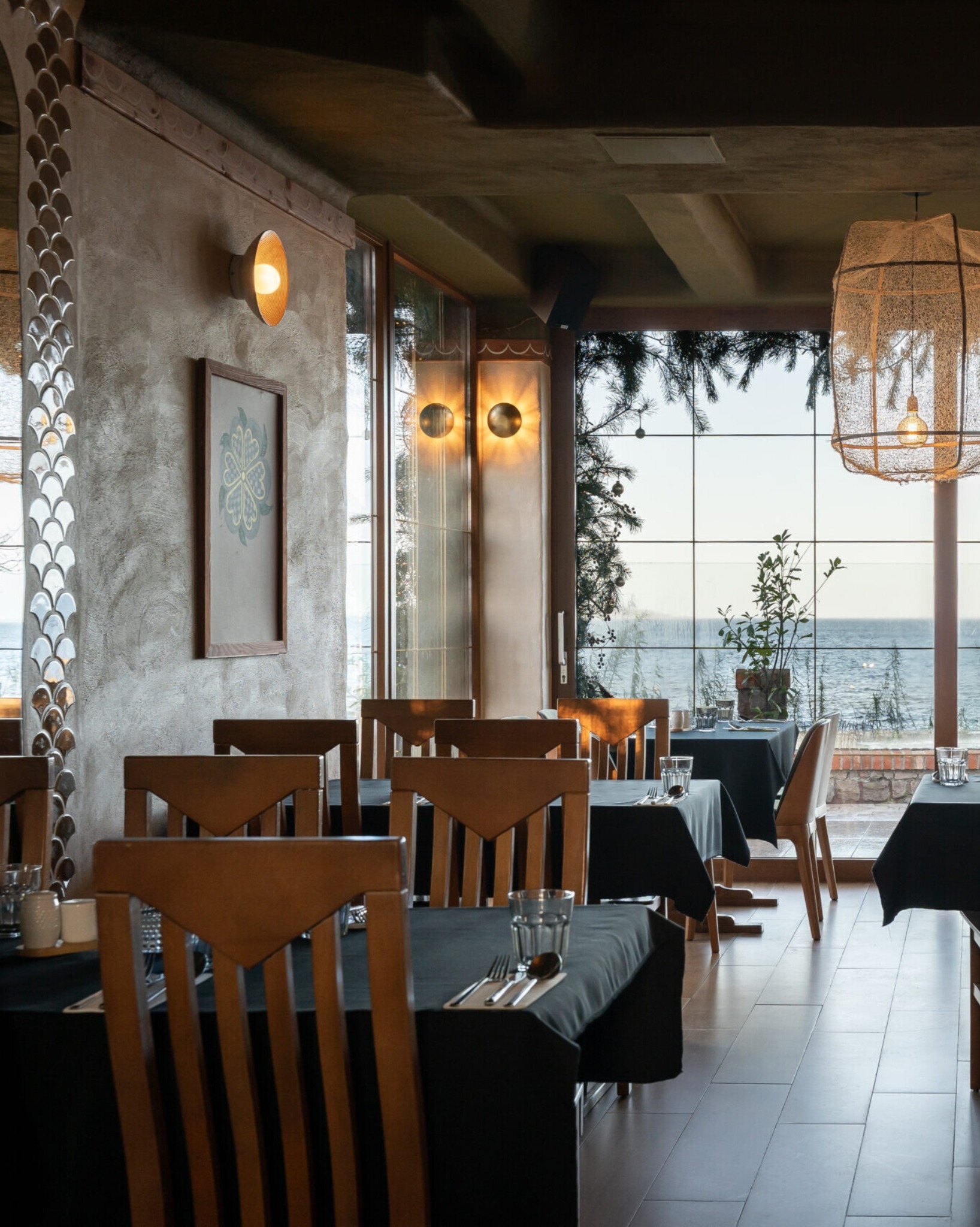 Wnętrze restauracji z widokiem na morze: drewniane krzesła, stoliki z czarnymi obrusami, dekoracyjne lampy i okno z widokiem na morze. Przytulna aranżacja.