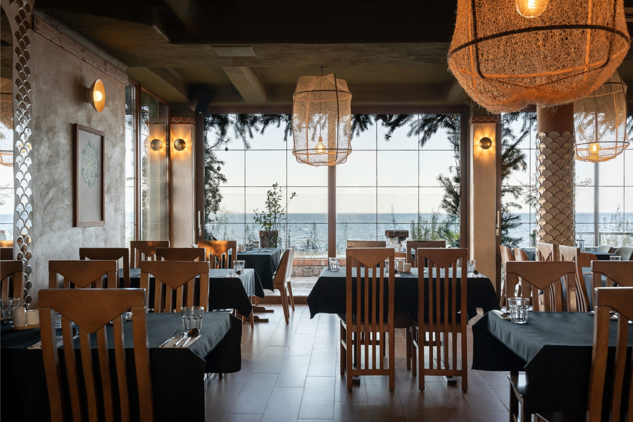 Elegancka restauracja z widokiem na morze. Drewniane krzesła i stoły z czarnymi obrusami. Dekoracyjne lampy wiszące. Duże okna z widokiem na błękitne morze.