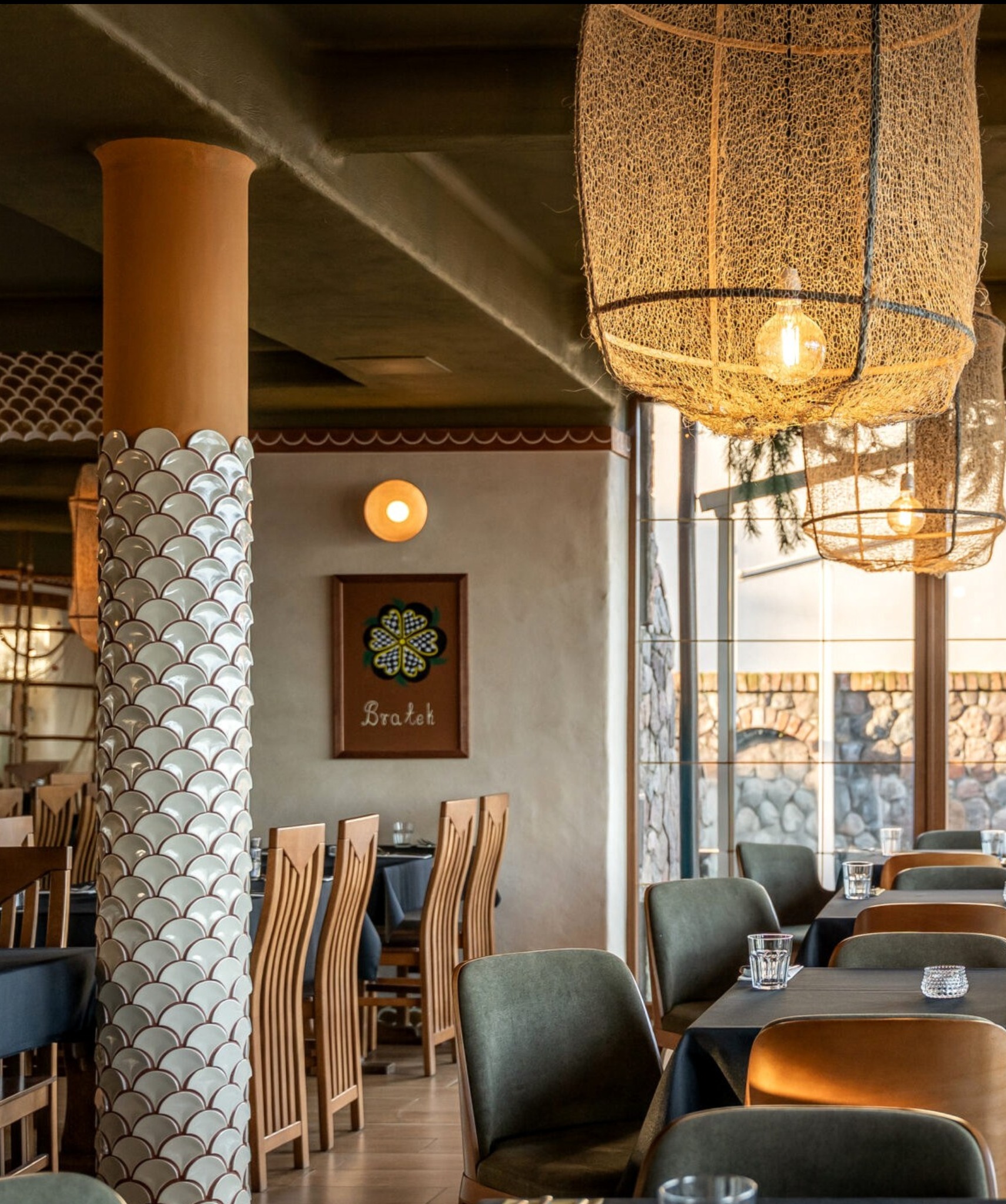 Wnętrze restauracji z kolumną zdobioną mozaiką w kształcie rybich łusek, wiszącymi lampami z naturalnego materiału i drewnianymi krzesłami przy nakrytych stołach.