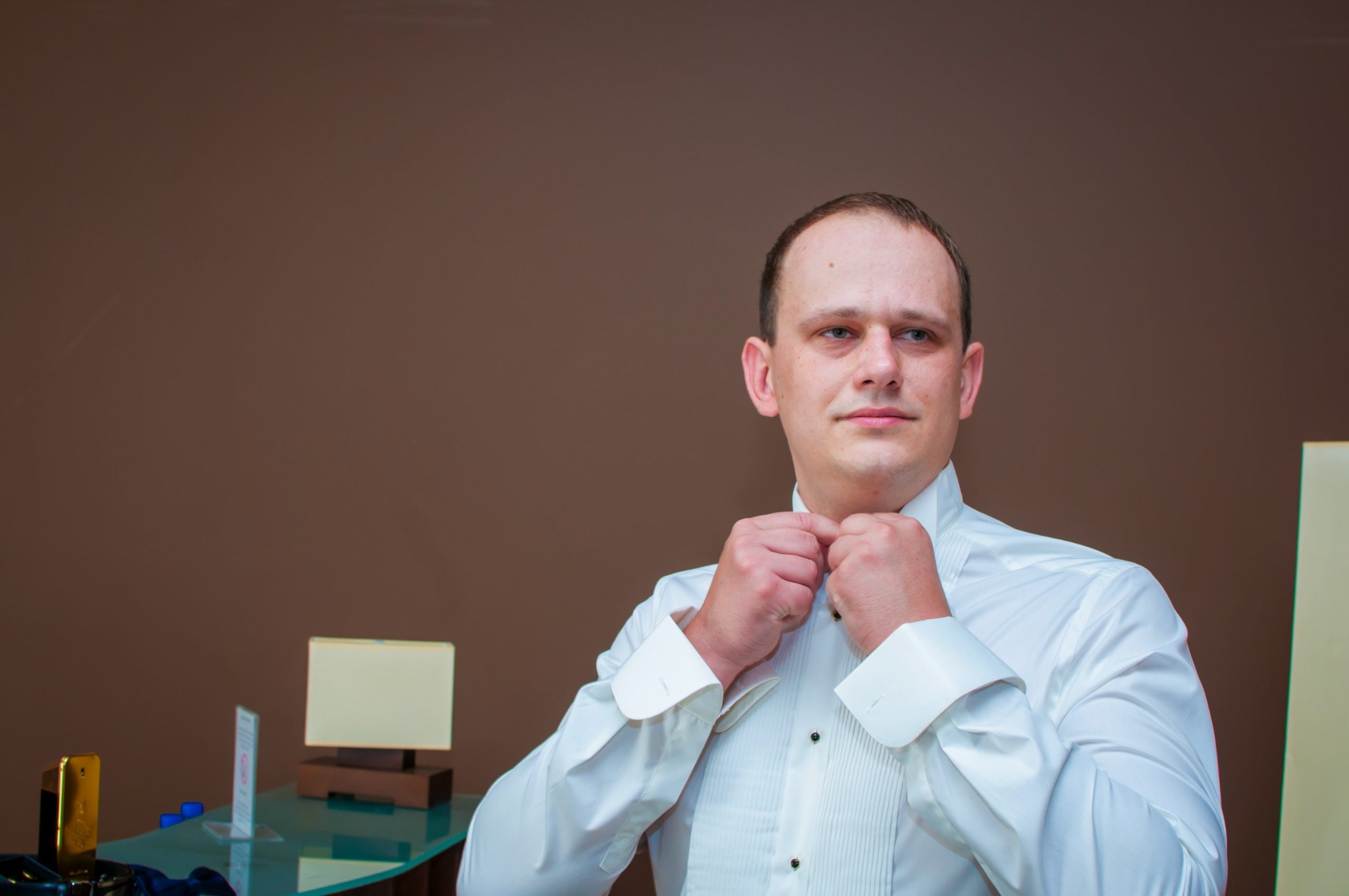 Mężczyzna w białej koszuli smokingowej zapinający guzik przy kołnierzyku, widoczny fragment szklanego stolika z akcesoriami