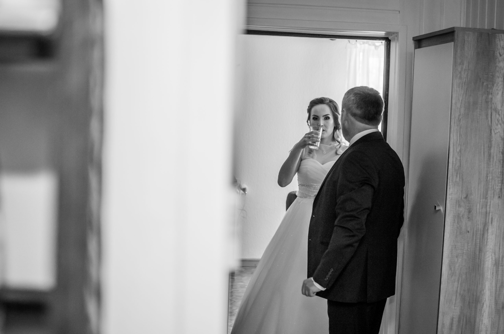 Czarno-białe zdjęcie panny młodej w sukni ślubnej, pijącej wodę z przezroczystej szklanki, z widocznym fragmentem sylwetki mężczyzny w garniturze