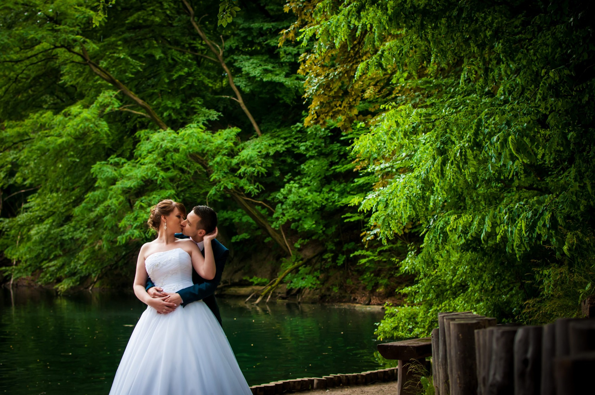 Panna młoda w białej sukni i pan młody w ciemnym garniturze całują się na tle zielonego lasu i jeziora.
