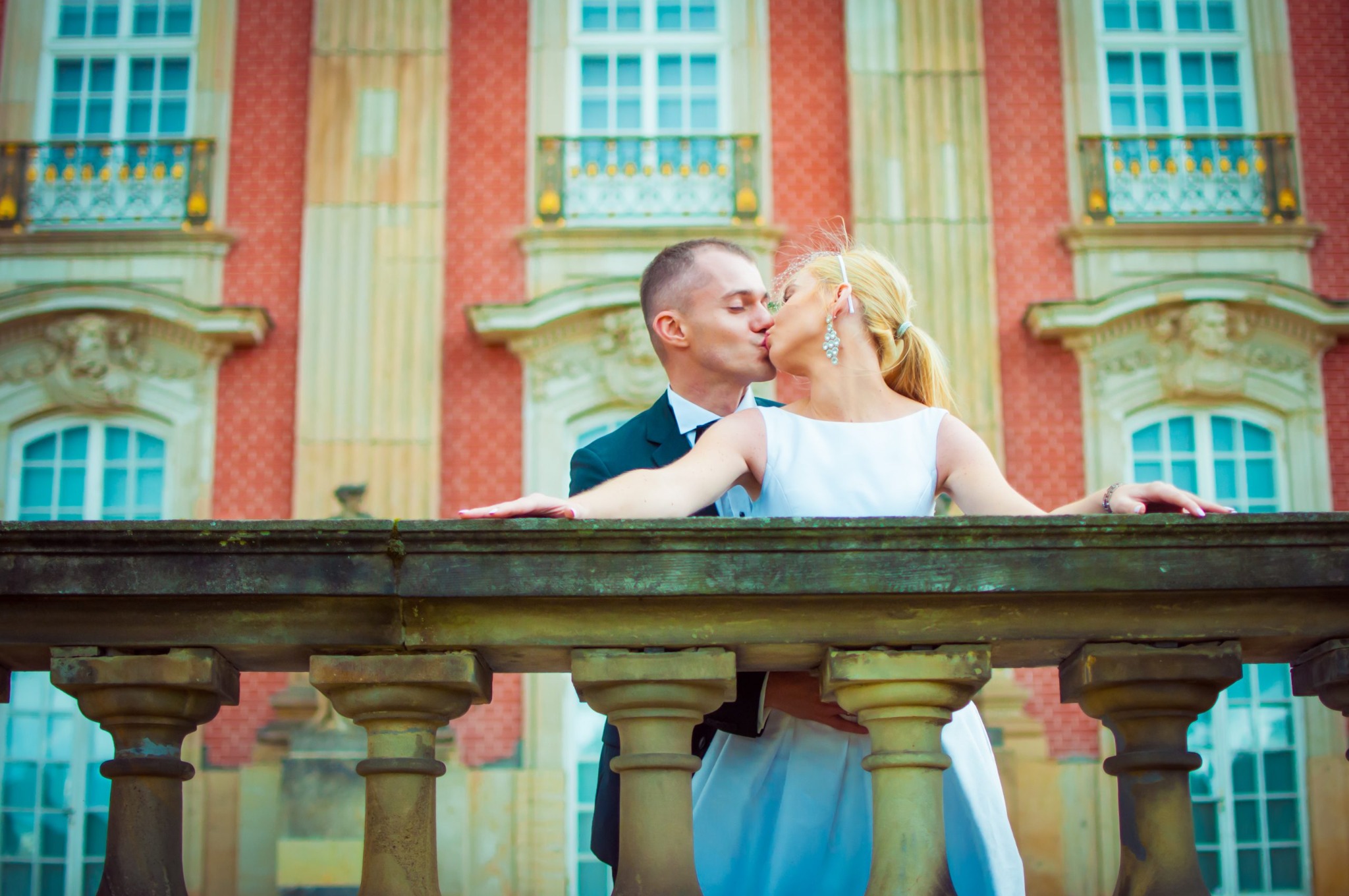 Pocałunek nowożeńców na tle pałacowej architektury z czerwonej cegły i białych okien, opierających się o kamienną balustradę z kolumnami.