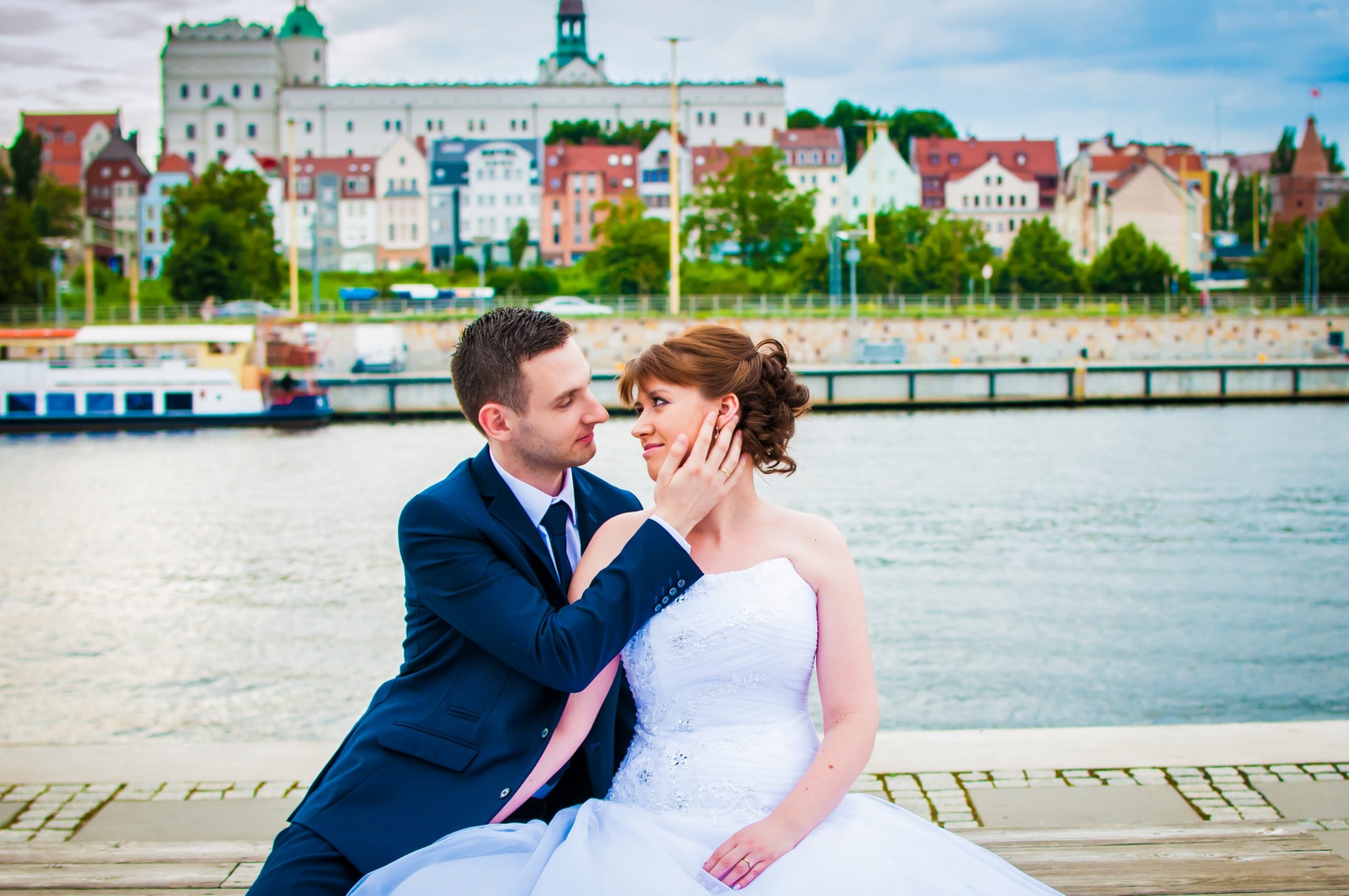 Para młoda siedząca na nabrzeżu z widokiem na miasto, on w granatowym garniturze dotyka jej twarzy, ona w białej sukni bez ramiączek, w tle rzeka i kolorowe budynki.