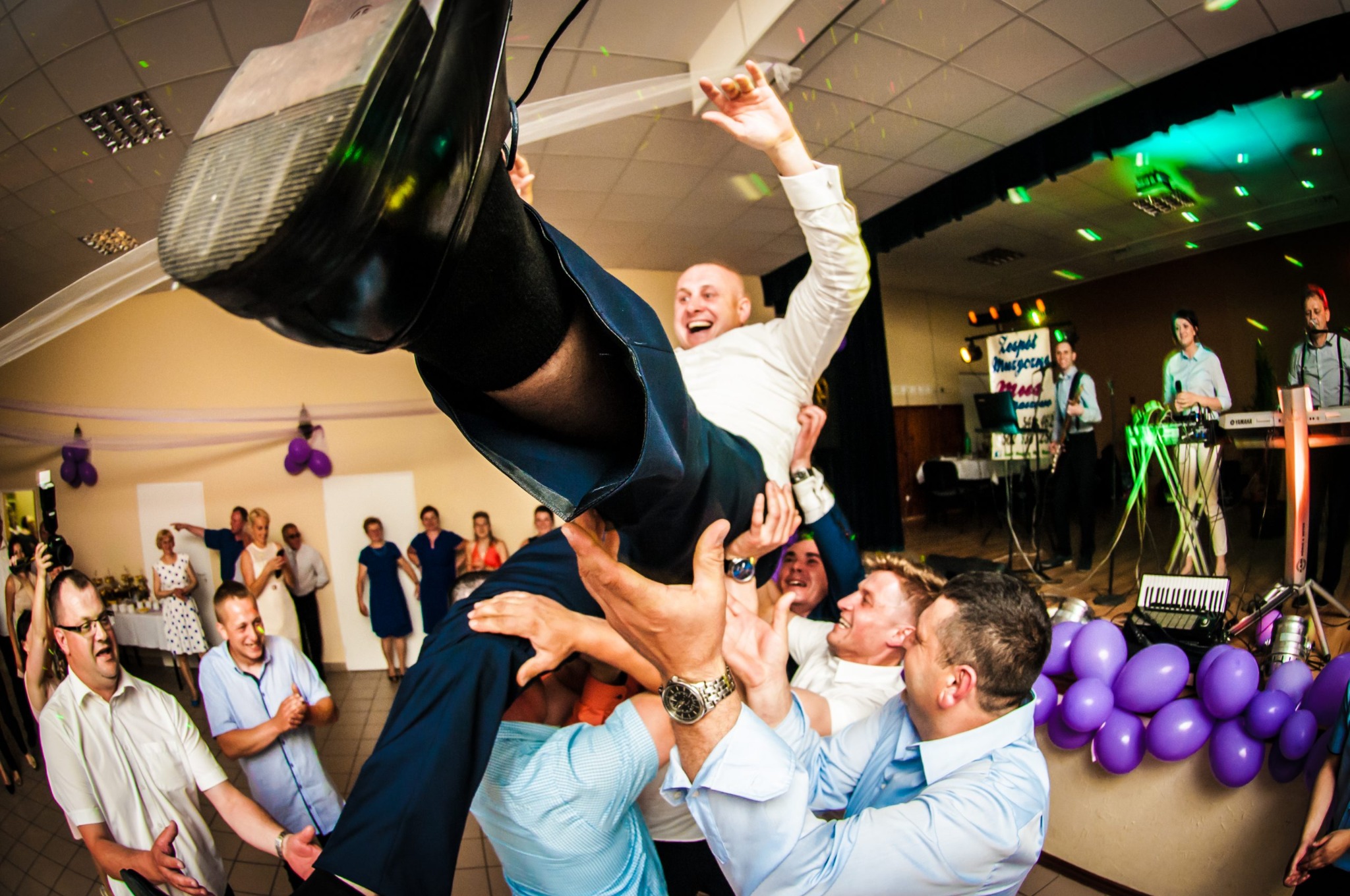 Uśmiechnięty pan młody wznoszony do góry przez gości weselnych podczas zabawy, w tle zespół muzyczny i dekoracje z fioletowych balonów.