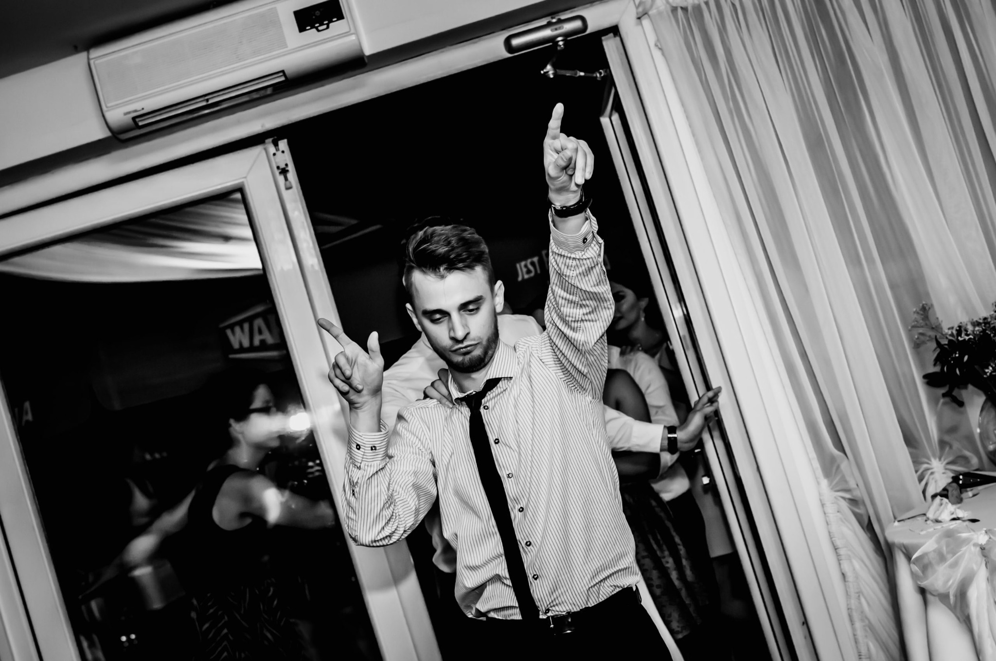 Mężczyzna w koszuli w paski i krawacie tańczy z uniesionymi rękami na czarno-białym zdjęciu z imprezy, widoczni inni goście i fragment dekoracji.