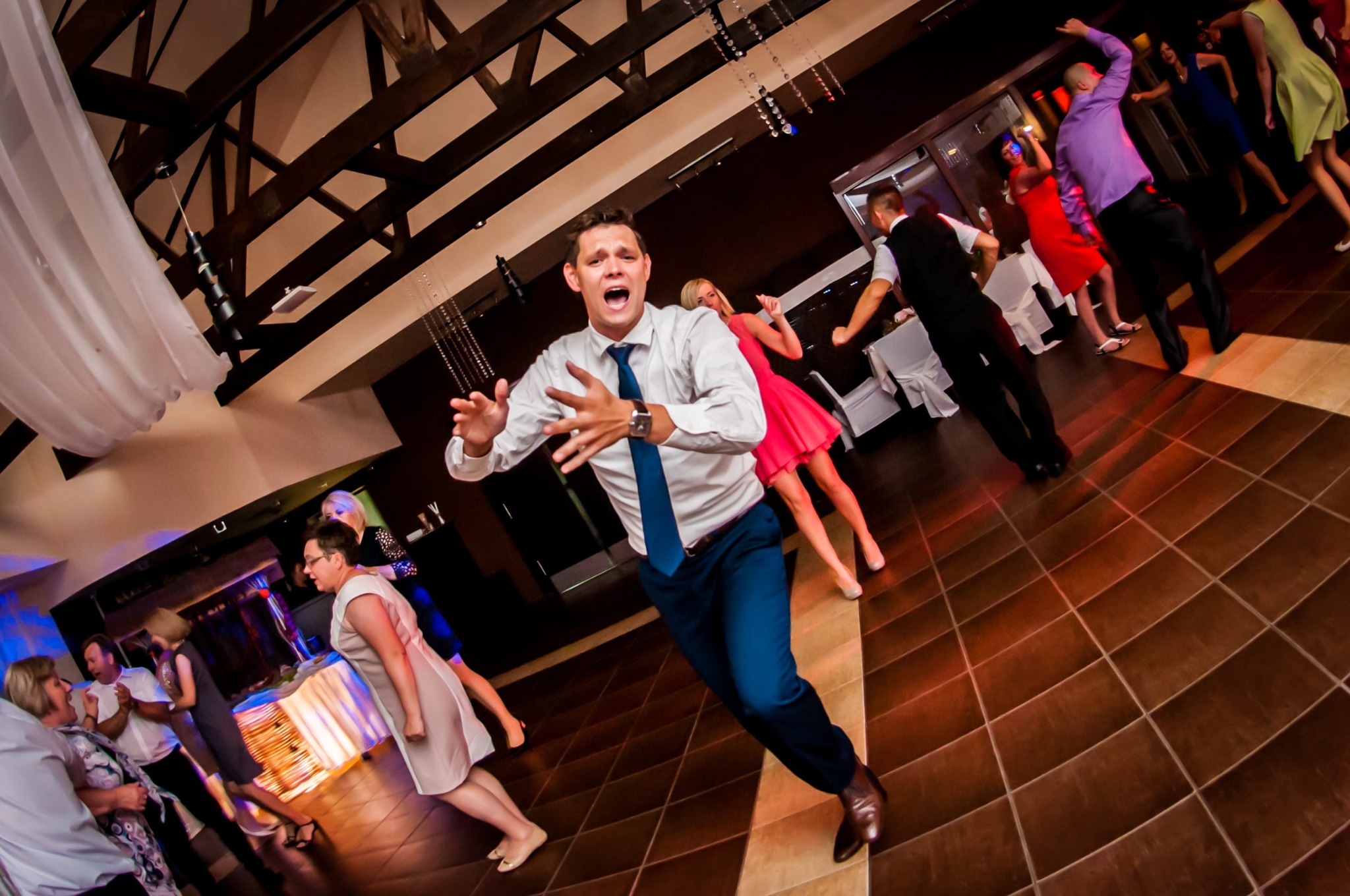 Rozentuzjazmowany mężczyzna w tańcu na parkiecie z brązowych płytek podczas imprezy, w tle bawiący się goście i dekoracje sali.