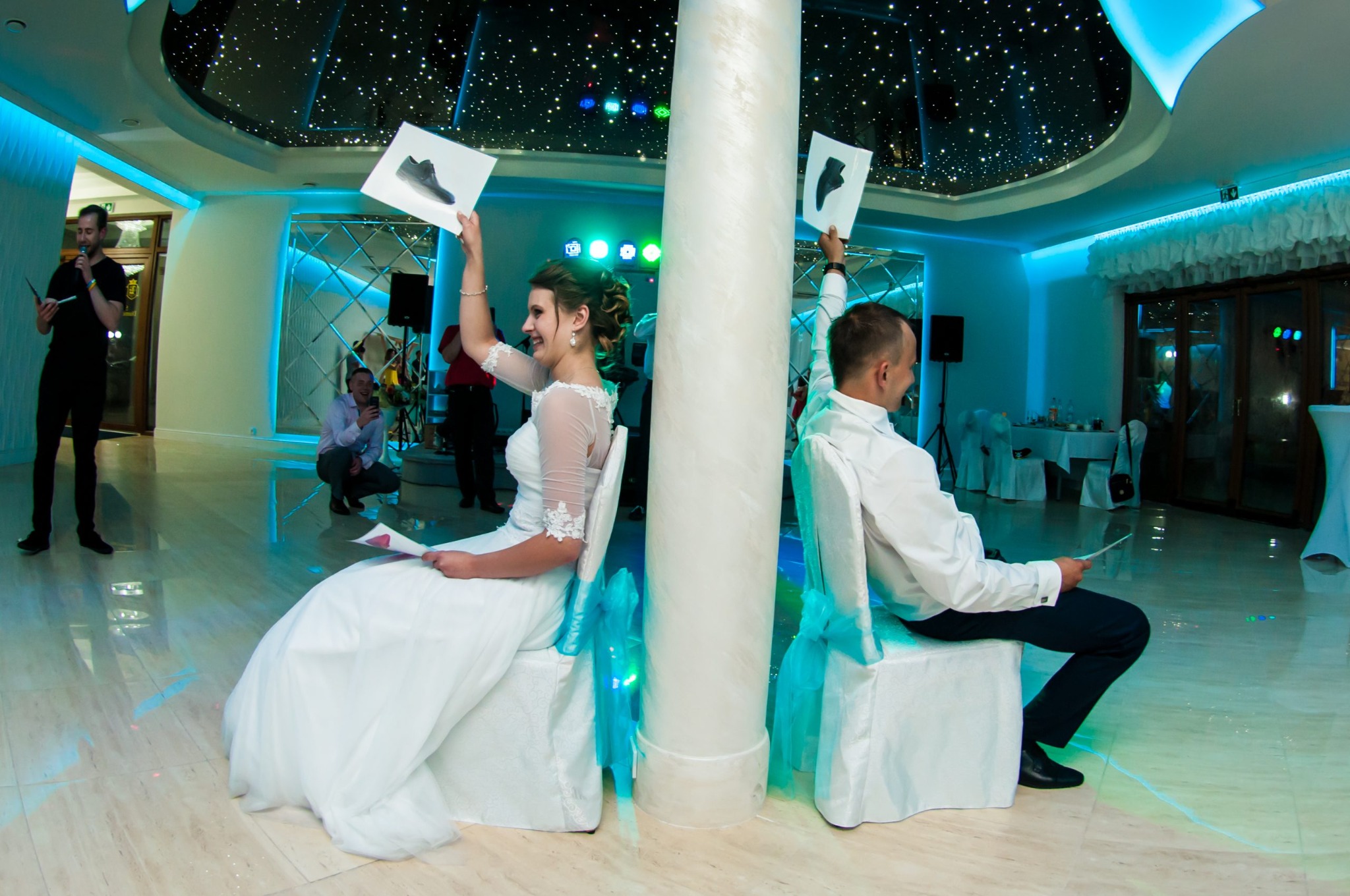 Para Młoda siedzi na krzesłach podczas weselnej zabawy z butami na kartkach, w tle oświetlona sala z dekoracją imitującą gwiazdy na suficie.