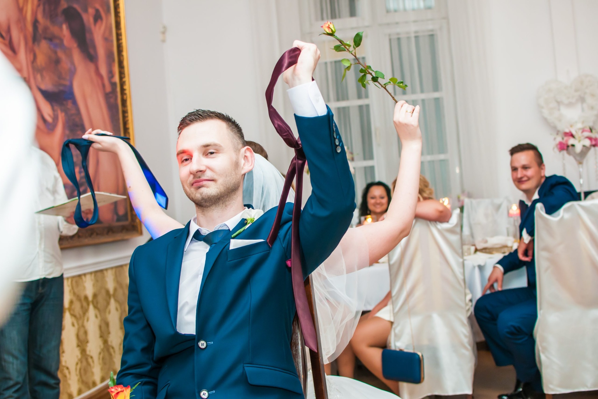Uśmiechnięty mężczyzna w niebieskim garniturze trzyma w górze krawat i różyczkę podczas oczepin na weselu, w tle goście i dekoracje.