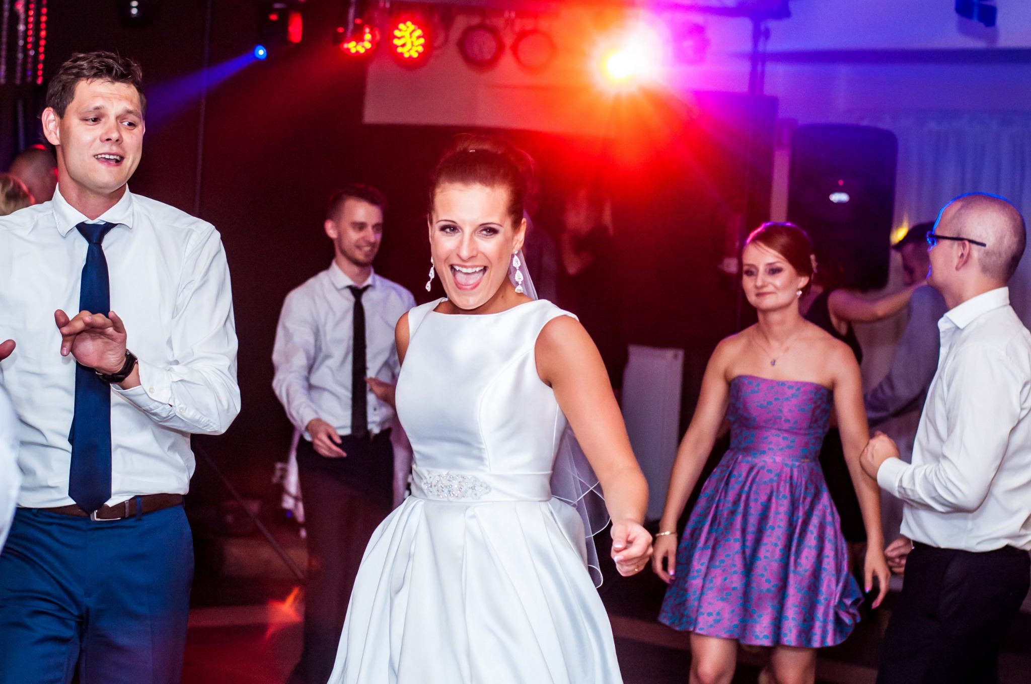 Rozpromieniona panna młoda w białej sukni tanecznej z ozdobnym pasem, w otoczeniu gości weselnych i kolorowych świateł dyskotekowych.