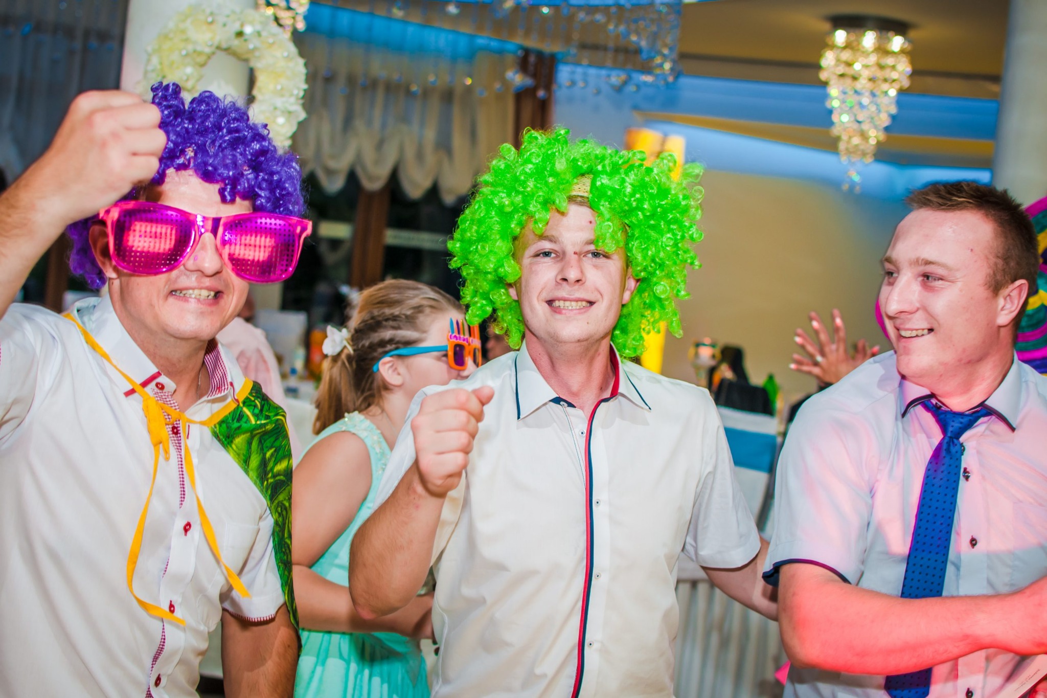 Trzech mężczyzn w perukach i zabawnych okularach pozuje podczas imprezy, w tle dekoracje i oświetlenie.