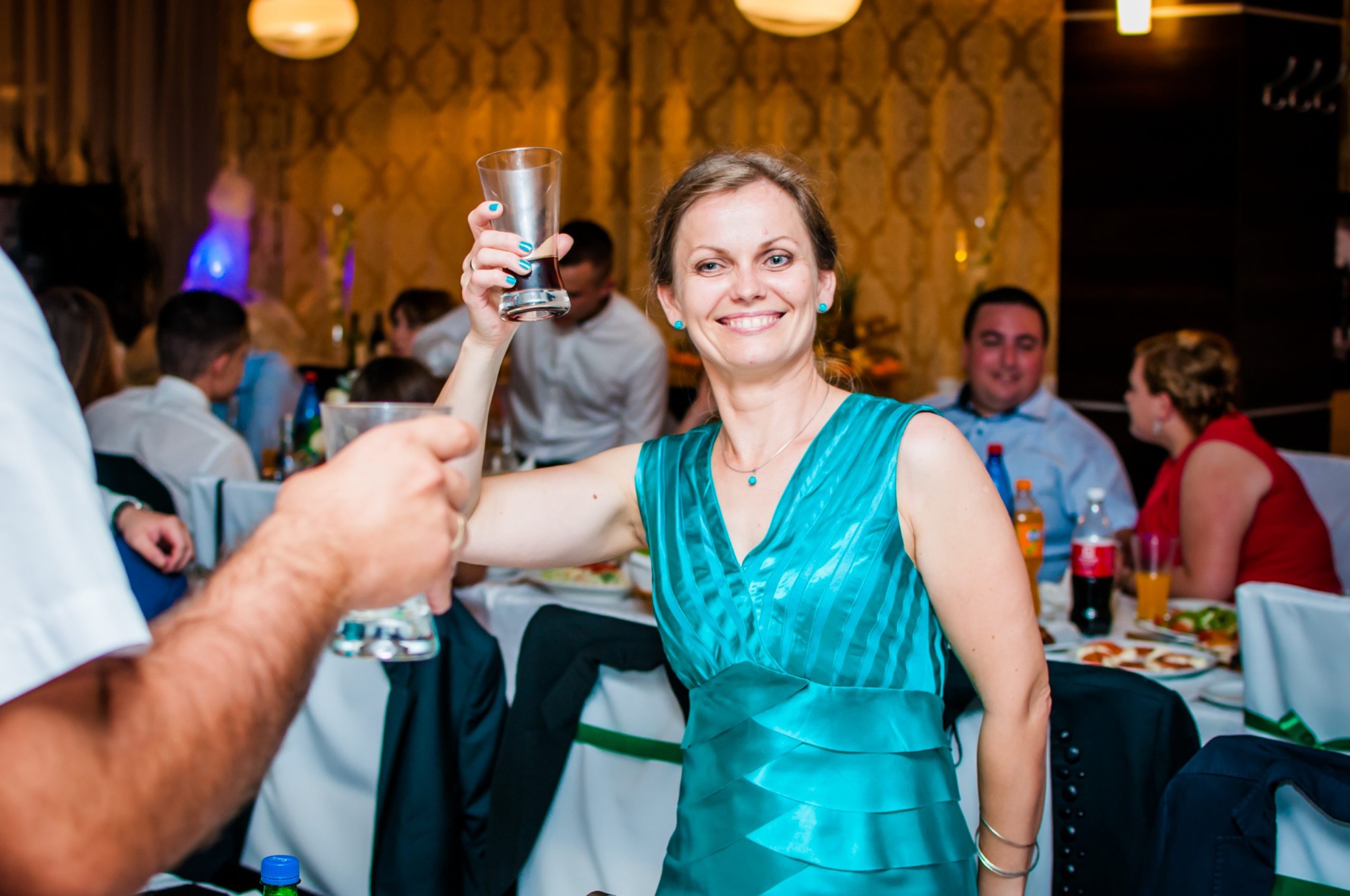 Uśmiechnięta kobieta w turkusowej sukience wznosi toast szklanką w dłoni podczas uroczystości, widoczni goście i dekoracje w tle.