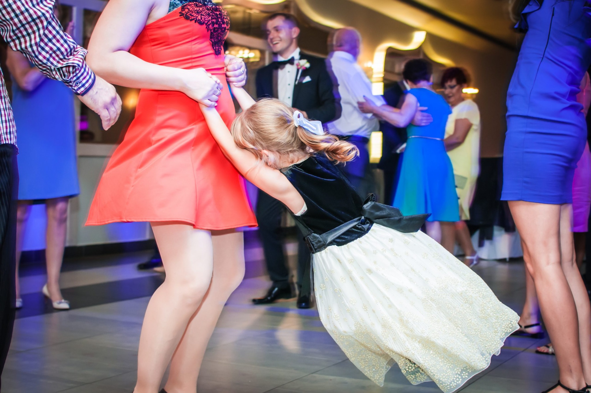Mała dziewczynka w sukience z tiulem przytulona do nogi kobiety w czerwonej sukience podczas tańca na parkiecie, w tle bawiący się goście weselni.