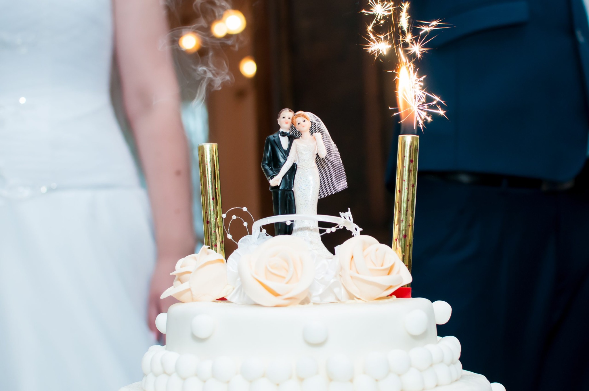Tort weselny z figurkami pary młodej, ozdobiony różami i płonącymi zimnymi ogniami, na tle fragmentów strojów gości.