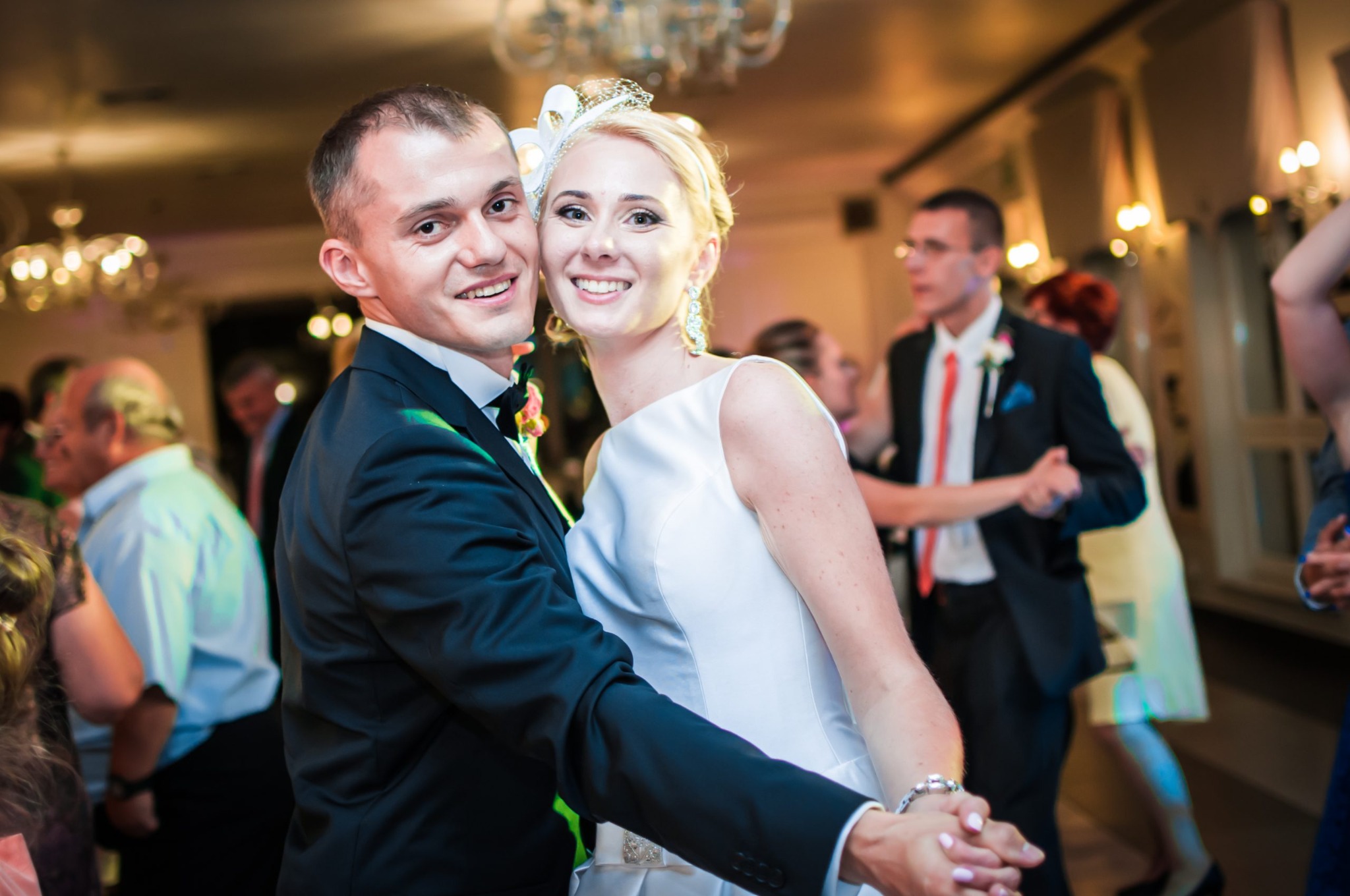 Uśmiechnięta para młoda w tańcu, on w czarnym smokingu z muszką, ona w białej sukni z ozdobą we włosach, na parkiecie inni goście weselni, ciepłe oświetlenie sali balowej.