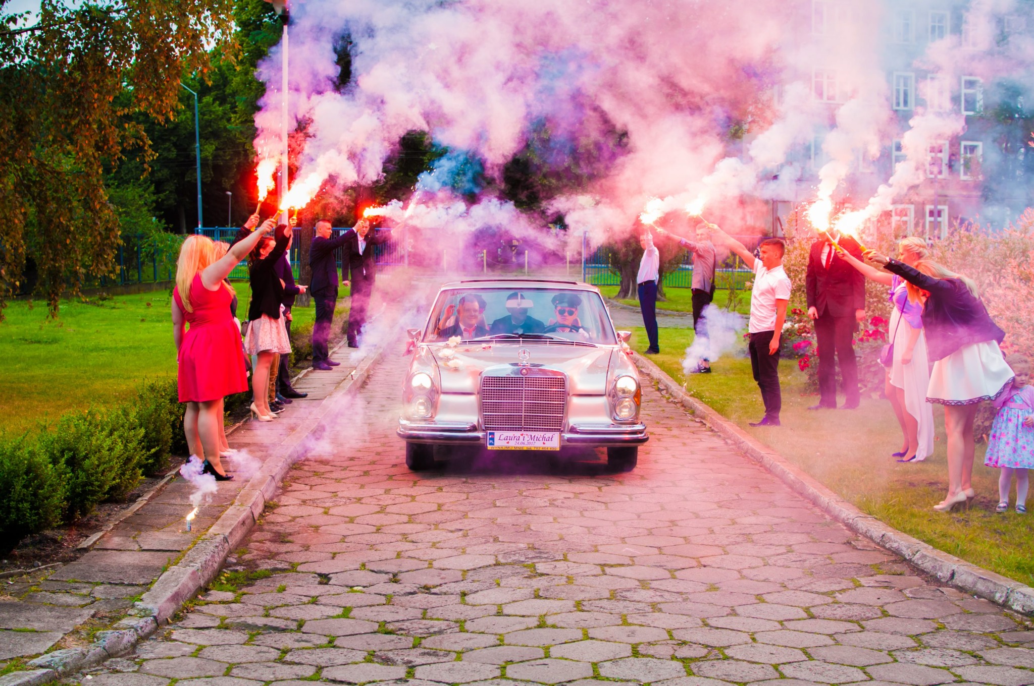 Nowożeńcy w srebrnym Mercedesie wjeżdżają na ceremonię otoczeni gośćmi z racami dymnymi tworzącymi kolorową mgłę.