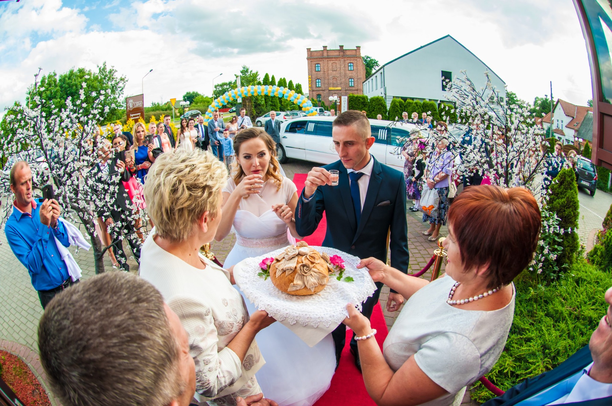 Młoda para wznosi toast wódką, trzymając kieliszki, podczas tradycyjnego powitania chlebem i solą na ślubie, z gośćmi i limuzyną w tle.