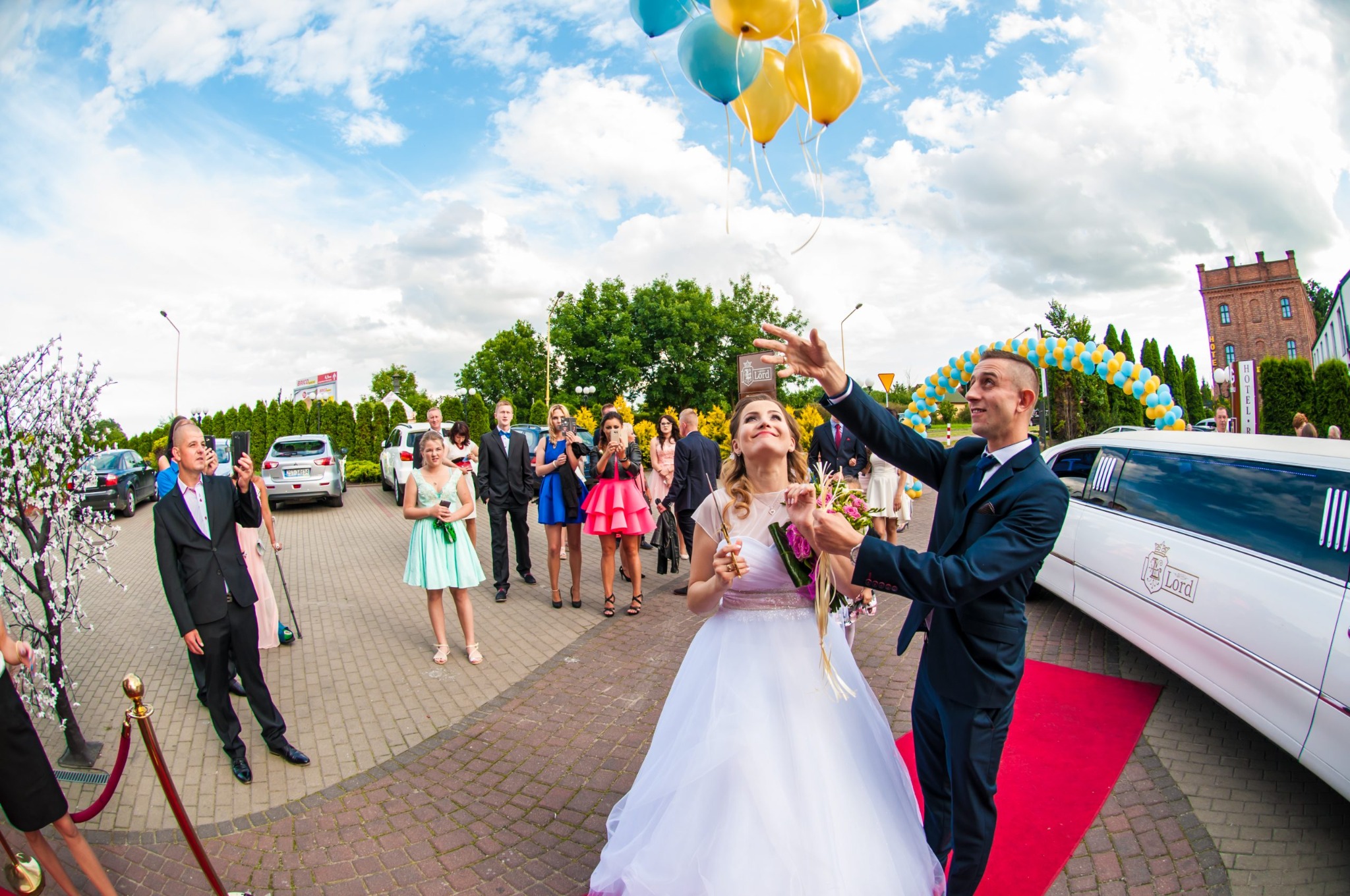 Panna młoda w białej sukni, z bukietem i balonami, stoi przed białą limuzyną z dekoracją balonową, w otoczeniu gości weselnych, fotografowanych przez inne osoby.