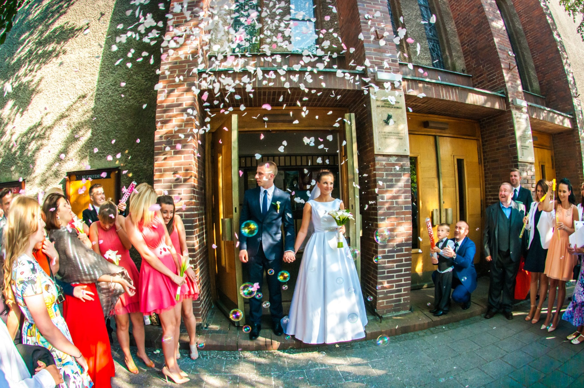 Para młoda wychodzi z kościoła, obsypywana konfetti i otoczona gośćmi, widoczne bańki mydlane w powietrzu, szeroki kąt ujęcia.