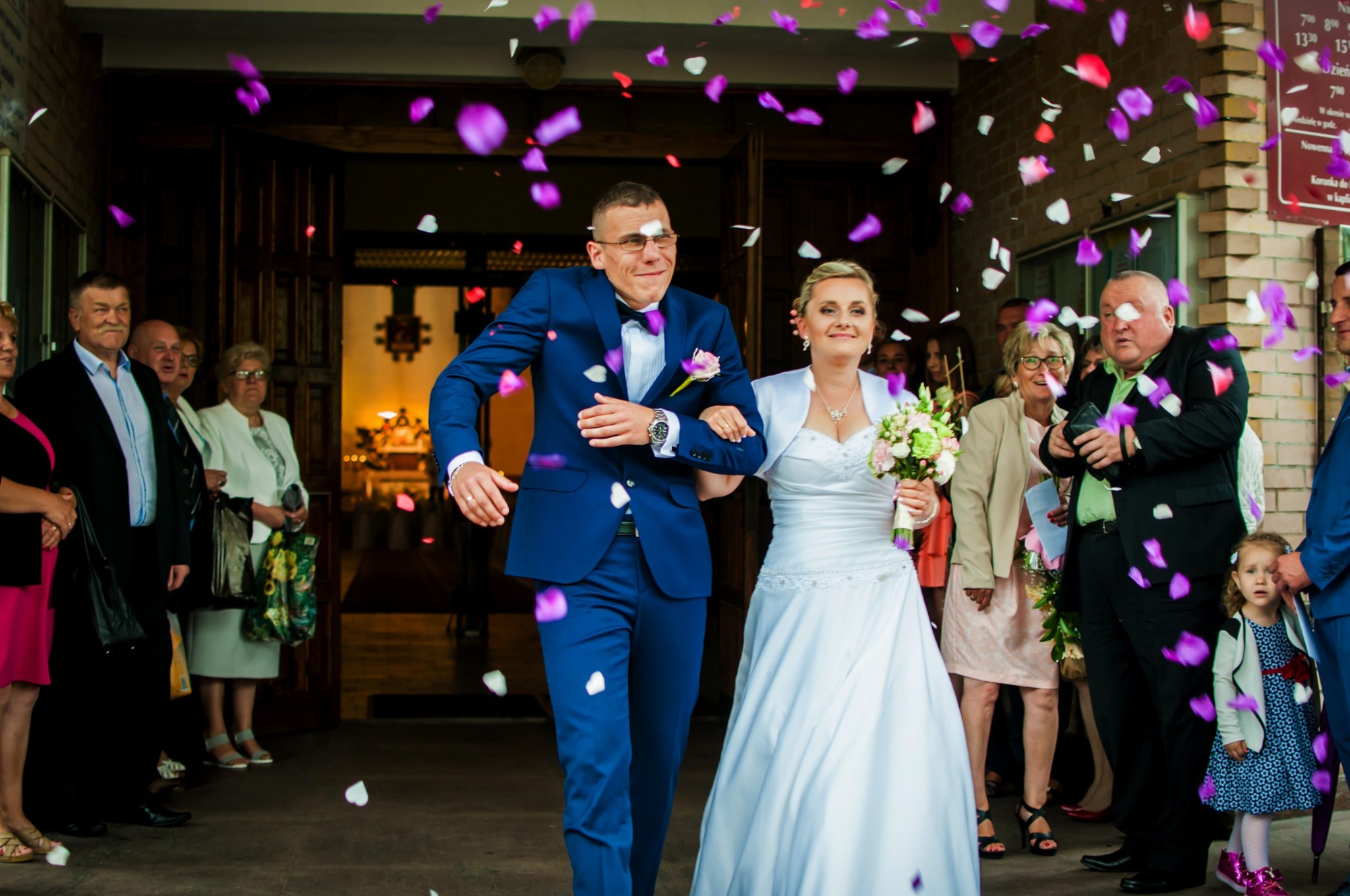 Para młoda w niebieskim garniturze i białej sukni wychodzi z kościoła, obsypywana konfetti z płatków kwiatów, w tle goście weselni.