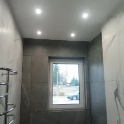 Wykończona łazienka z szarymi i białymi płytkami imitującymi marmur, widok na okno z widocznym samochodem na zewnątrz