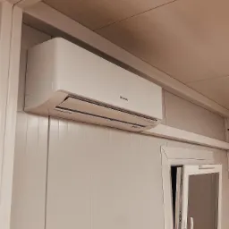 Biała jednostka klimatyzacyjna Hisense zamontowana pod sufitem w pomieszczeniu z panelowymi ścianami, widoczny fragment okna z uchylonym skrzydłem.